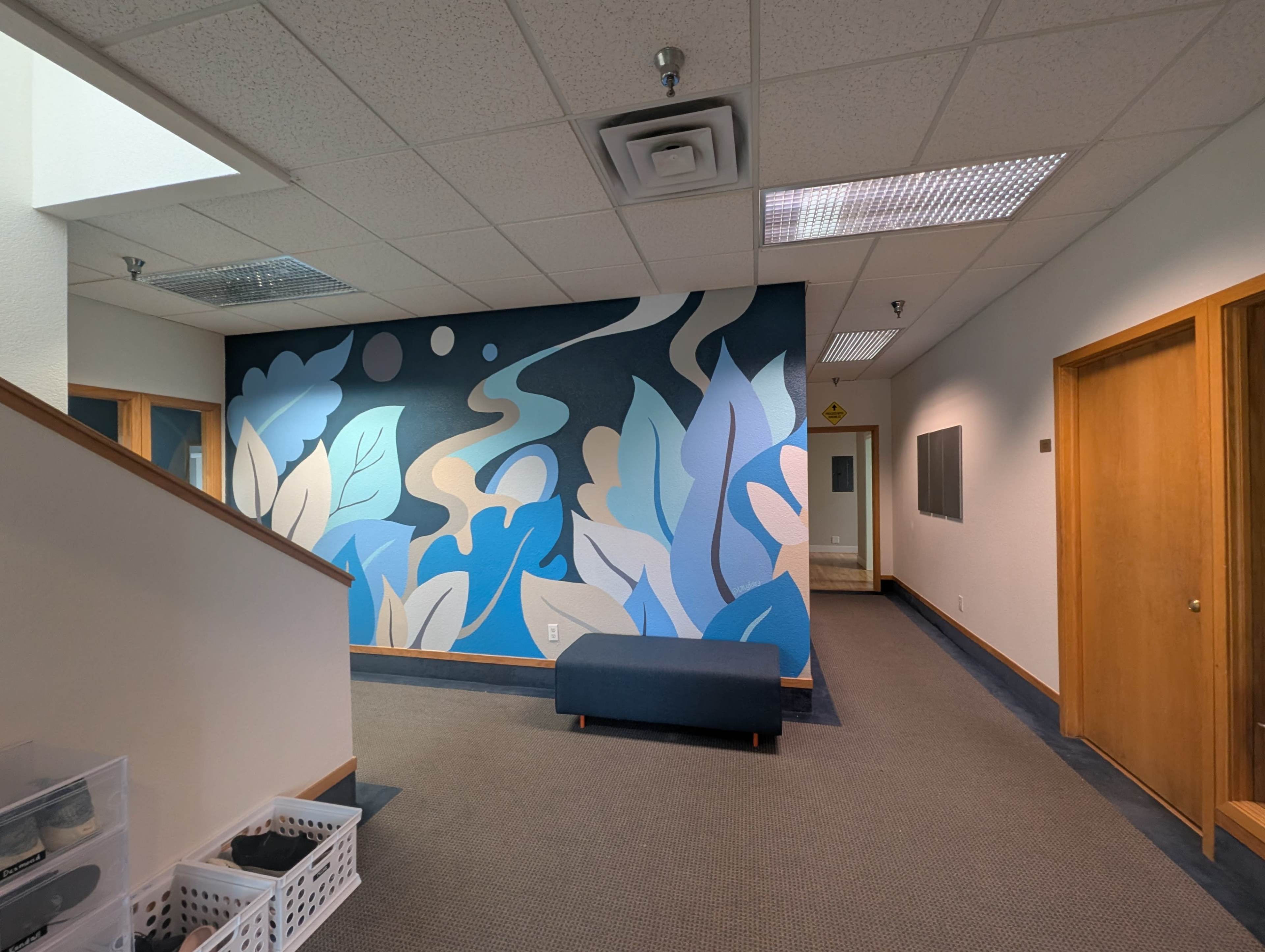 The image shows a hallway with a mural of abstract blue and beige shapes on one wall, a staircase, and several doorways leading to different rooms.