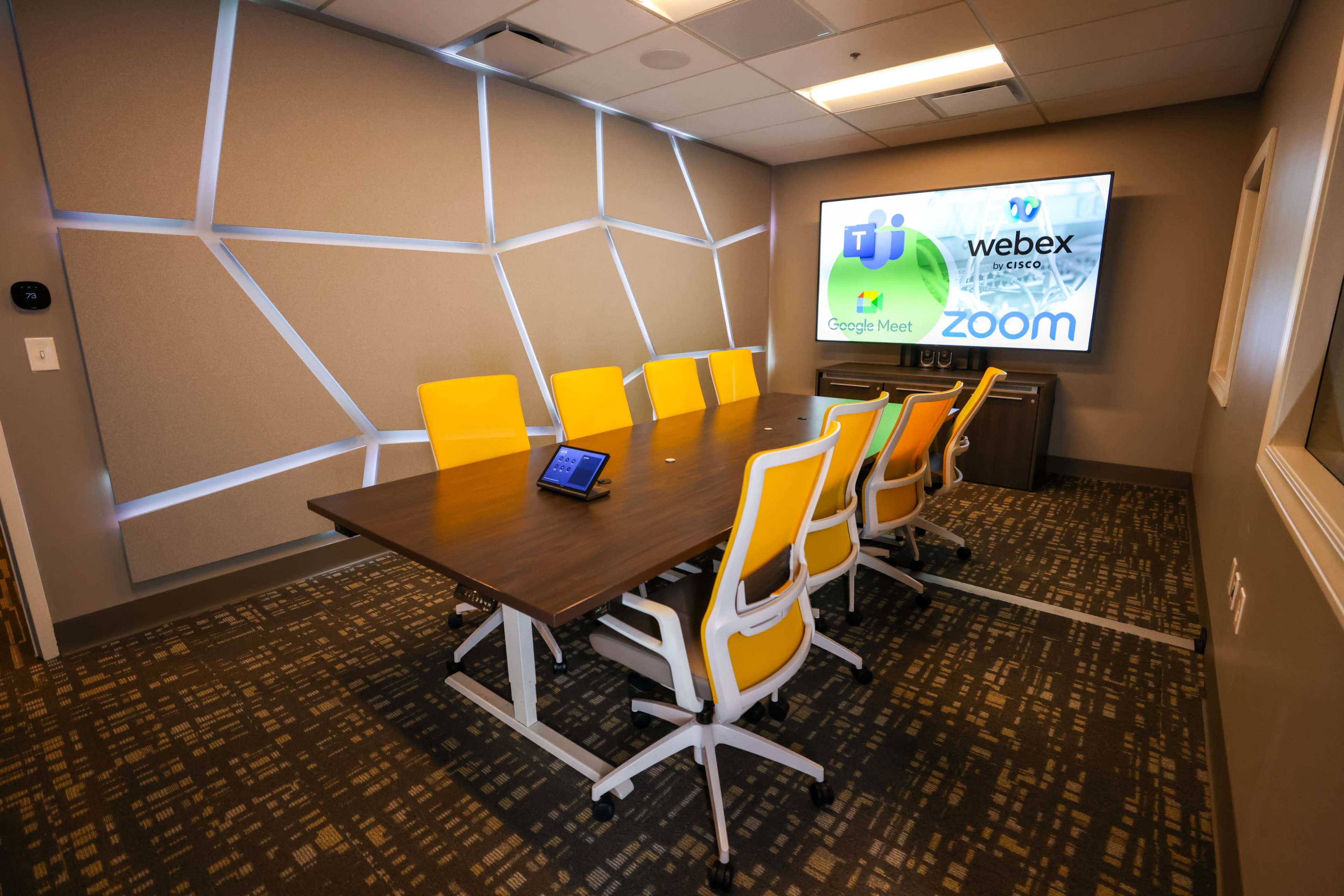 A modern conference room features a long wooden table with yellow chairs and a screen displaying logos for various video conferencing platforms.