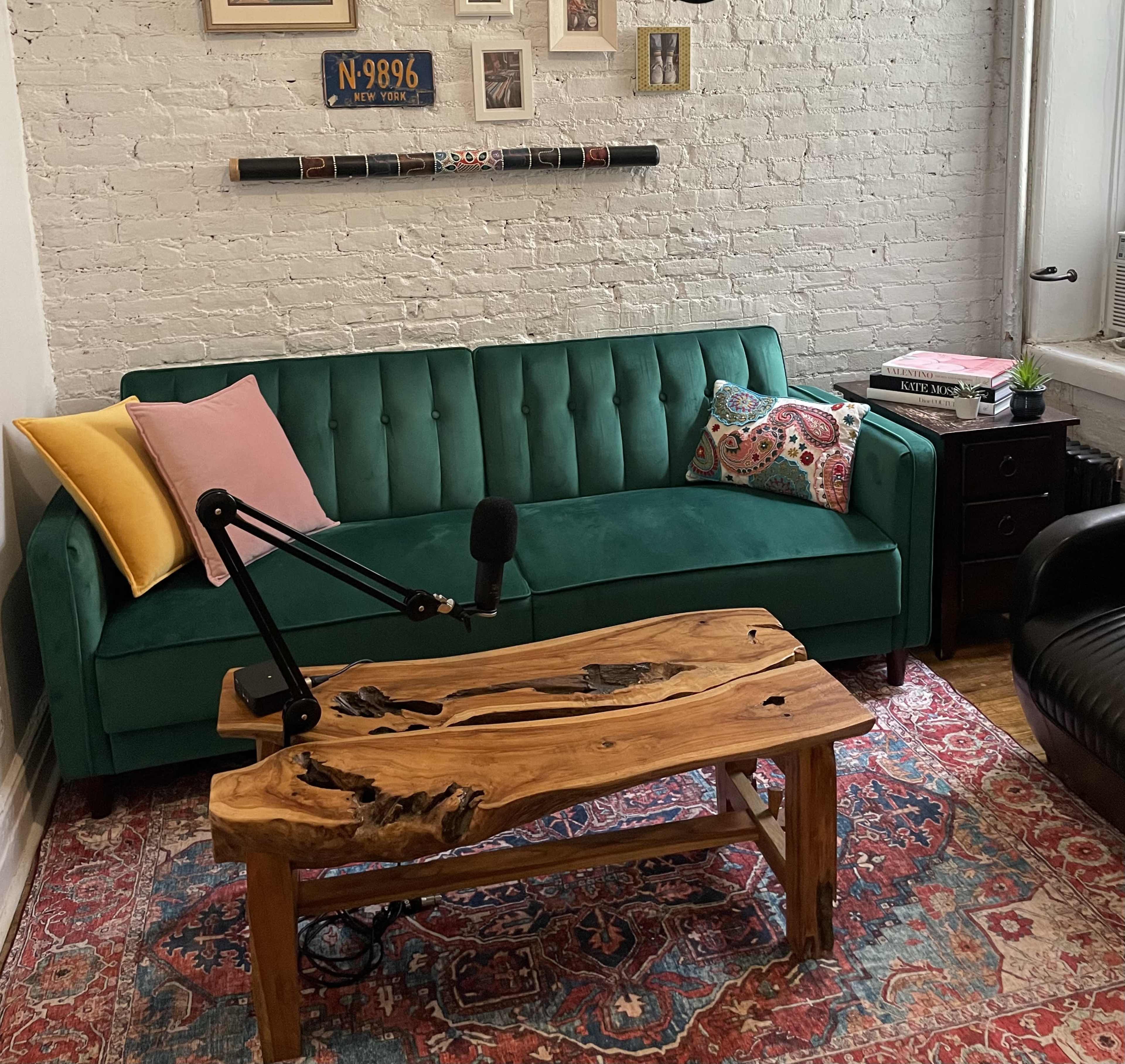 A green velvet sofa with colorful cushions faces a wooden coffee table on a patterned rug in a bright, minimalist room with exposed brick walls.