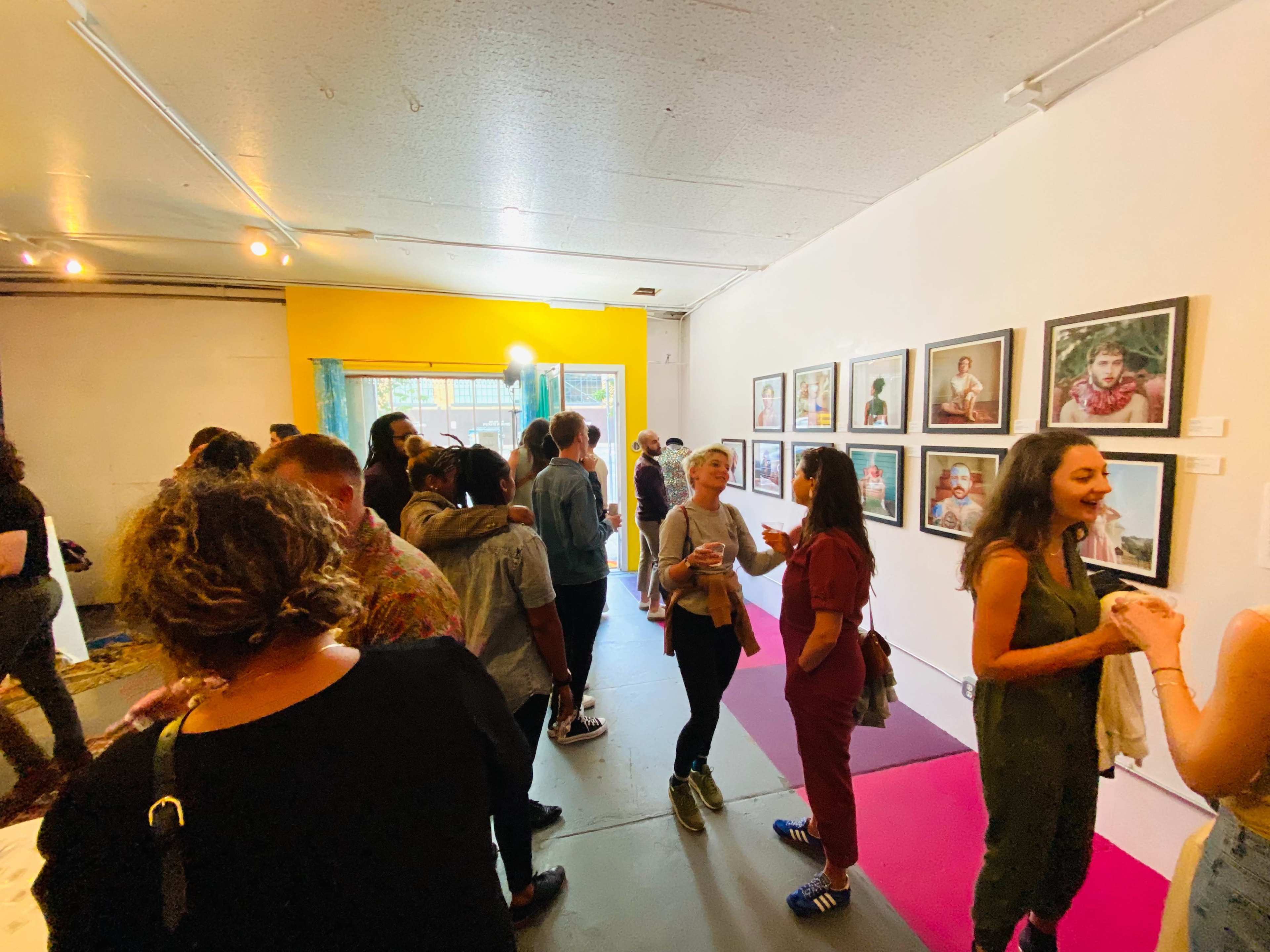 A crowded art gallery features attendees engaging with framed portraits on the walls.