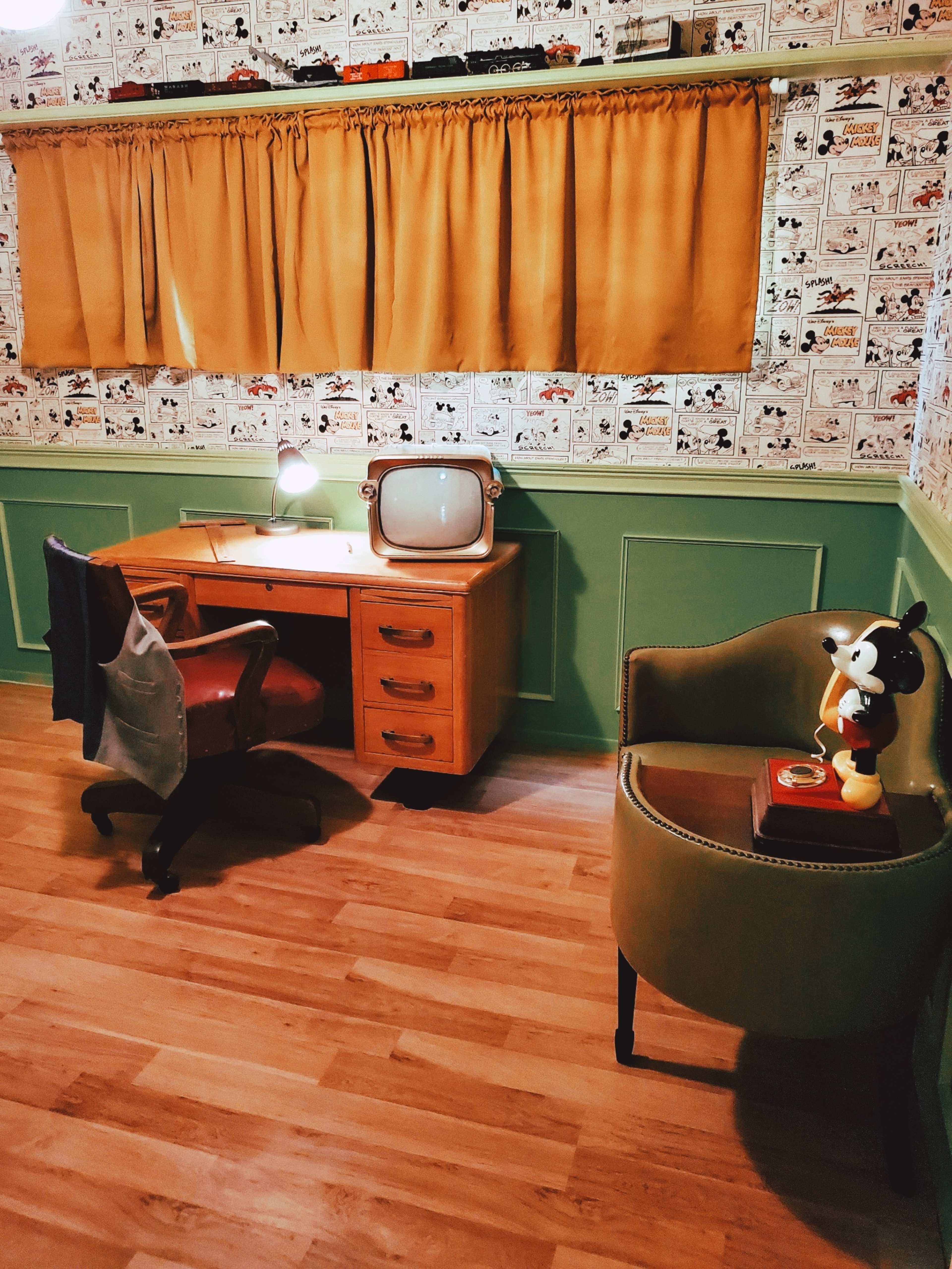 The room features a vintage desk with a lamp and an old television, alongside a green and yellow color scheme with a patterned wallpaper.
