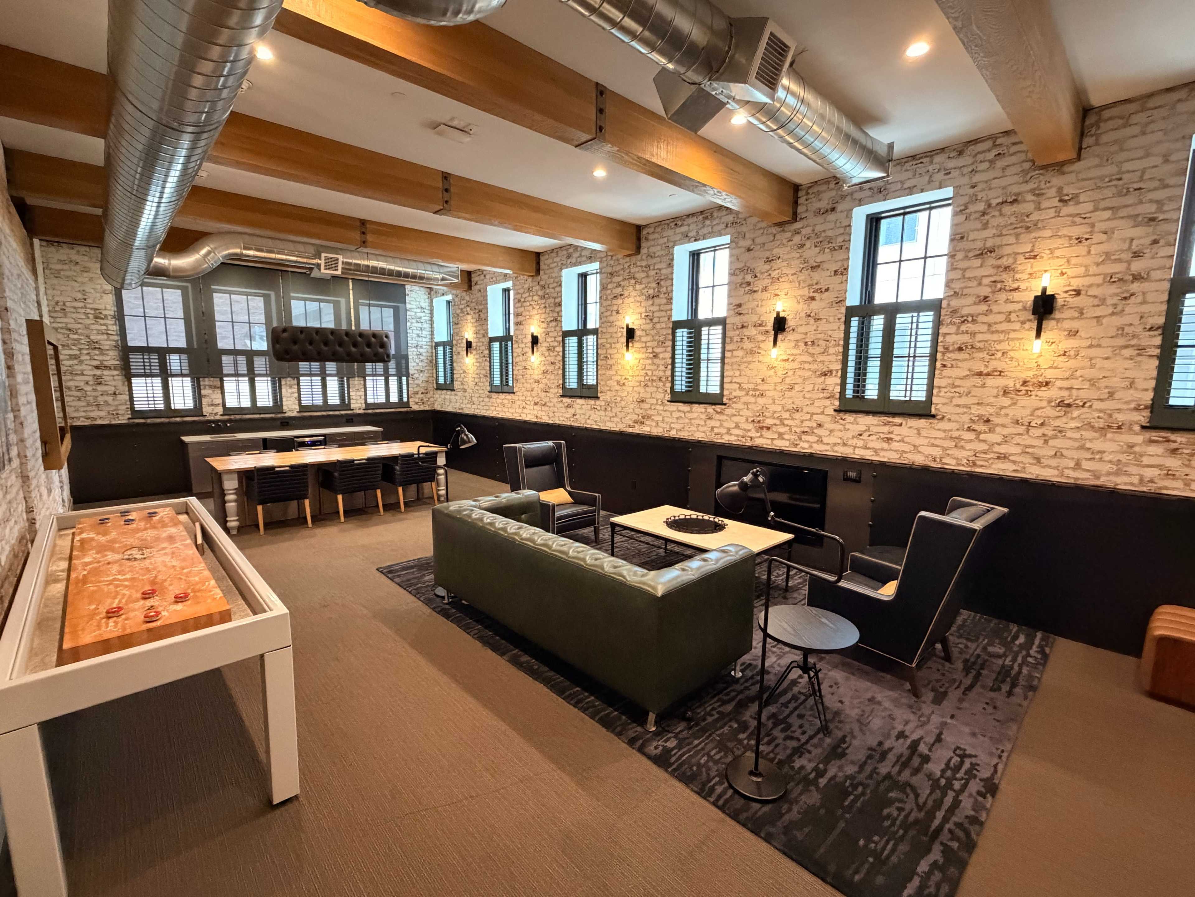 The image shows a spacious game room with a foosball table, comfortable seating, and large windows featuring a mix of brick and painted walls.