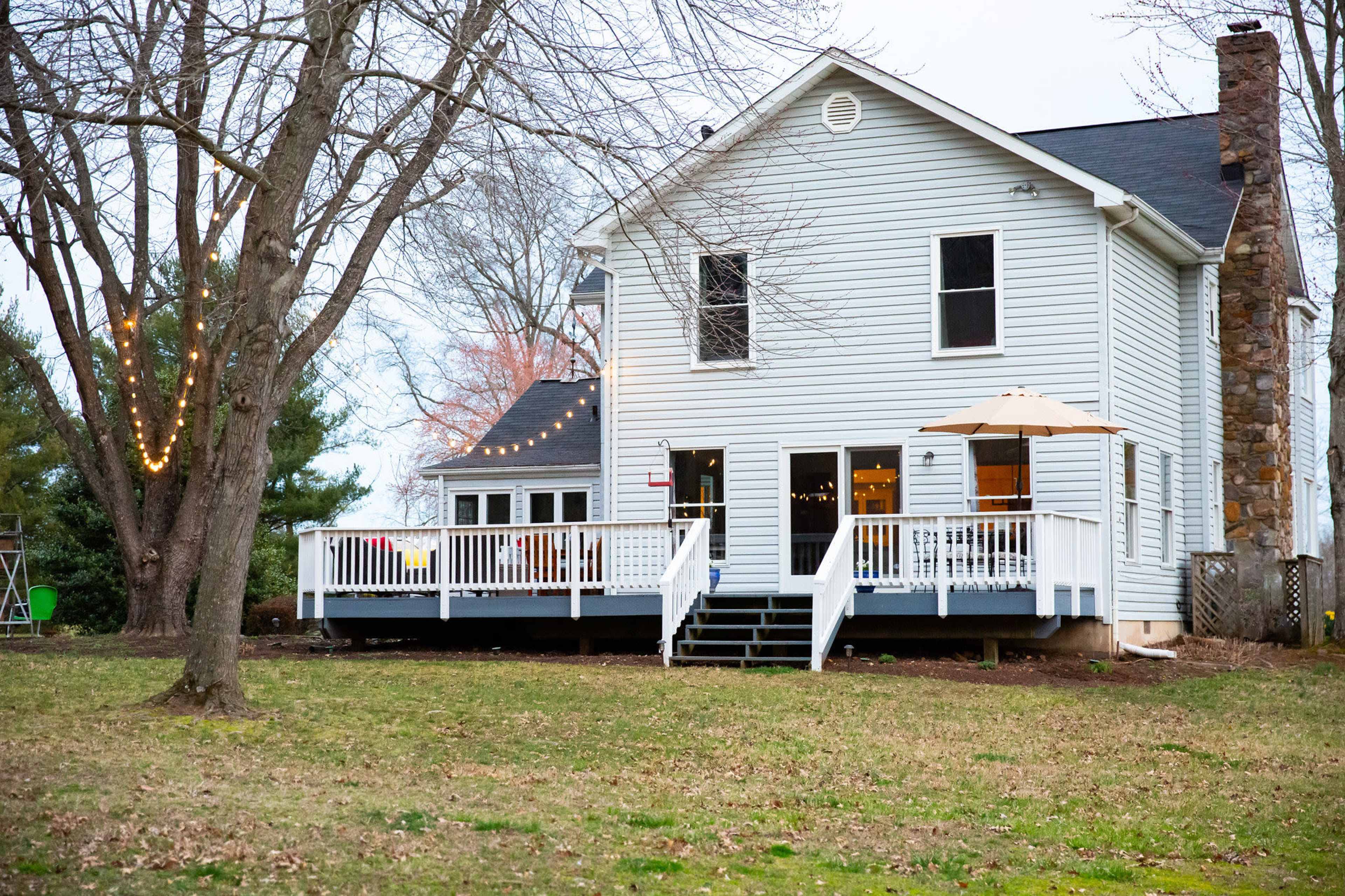 The image shows the back of a white two-story house with a deck, surrounded by trees in a grassy yard.