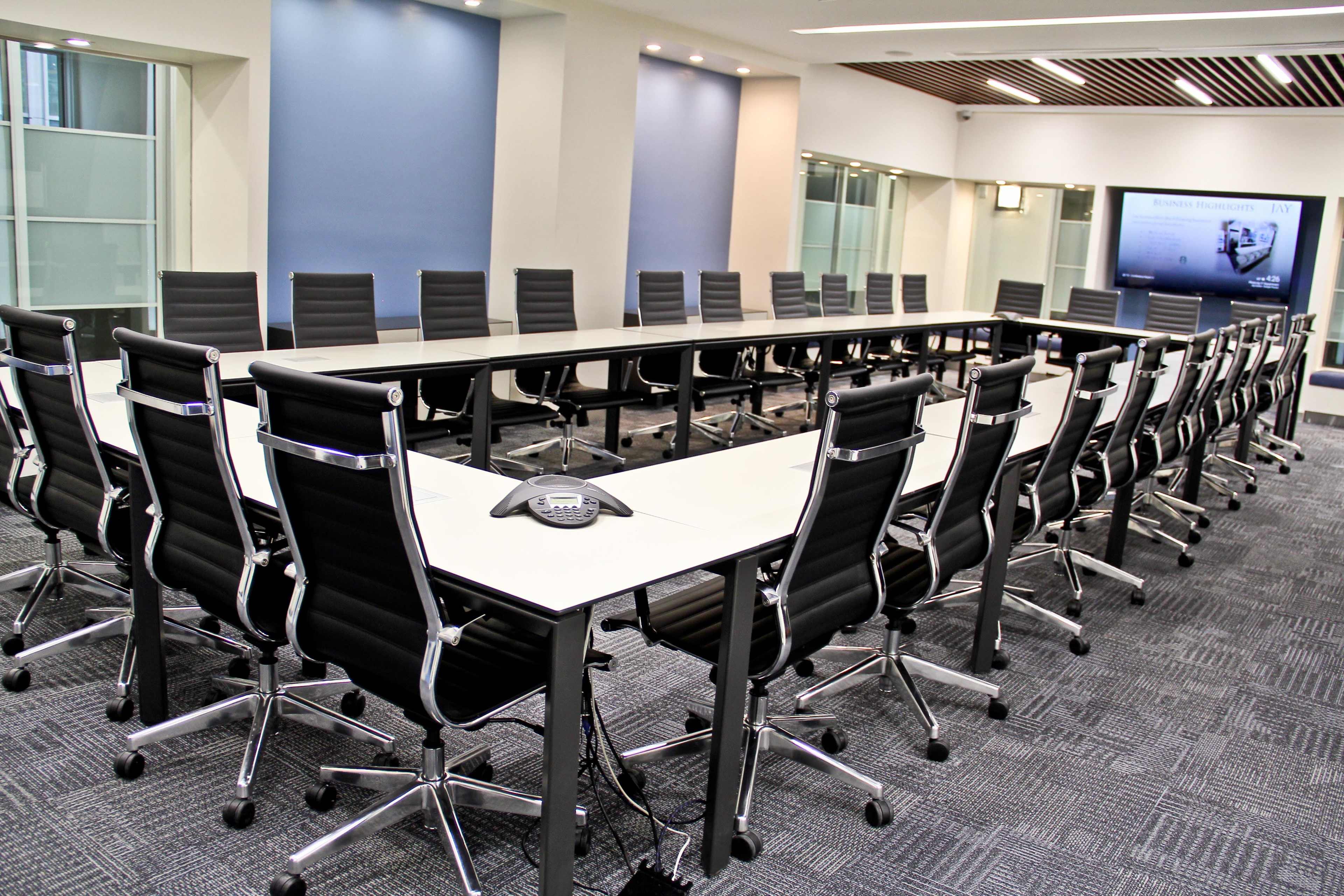 A large conference room features a long rectangular table surrounded by black ergonomic chairs and equipped with a speakerphone.