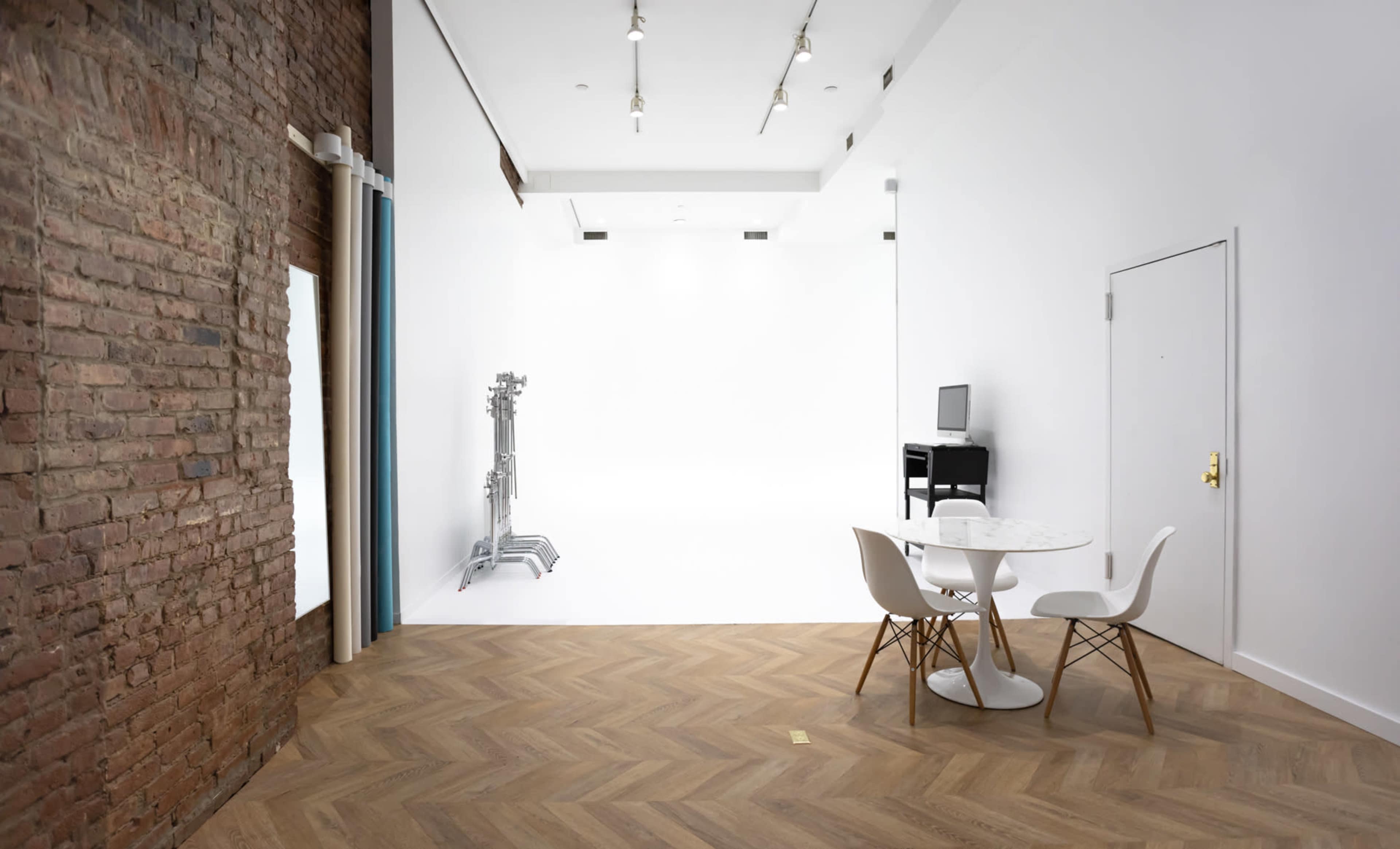 The image shows a modern interior space with a brick wall on one side, a white backdrop, a table with two chairs, and a small black cabinet.