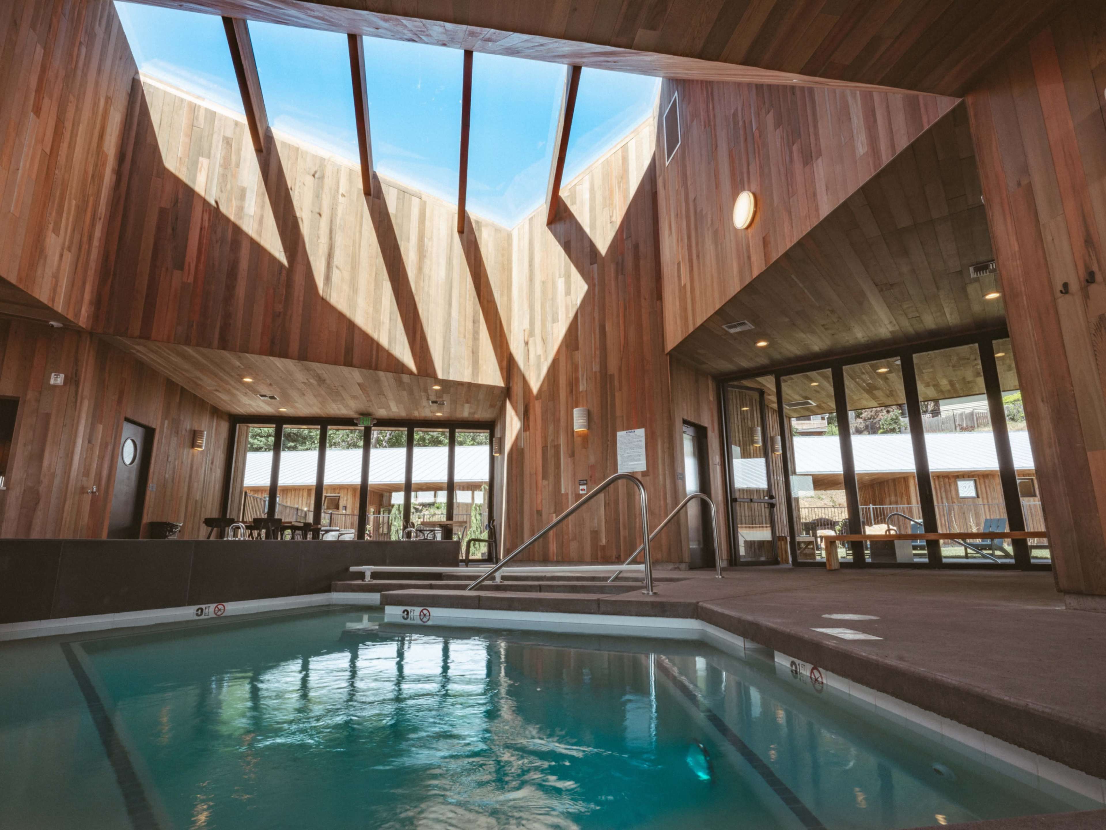 The image shows an indoor pool area with large wooden walls and a slanted ceiling that allows sunlight to stream in through skylights.