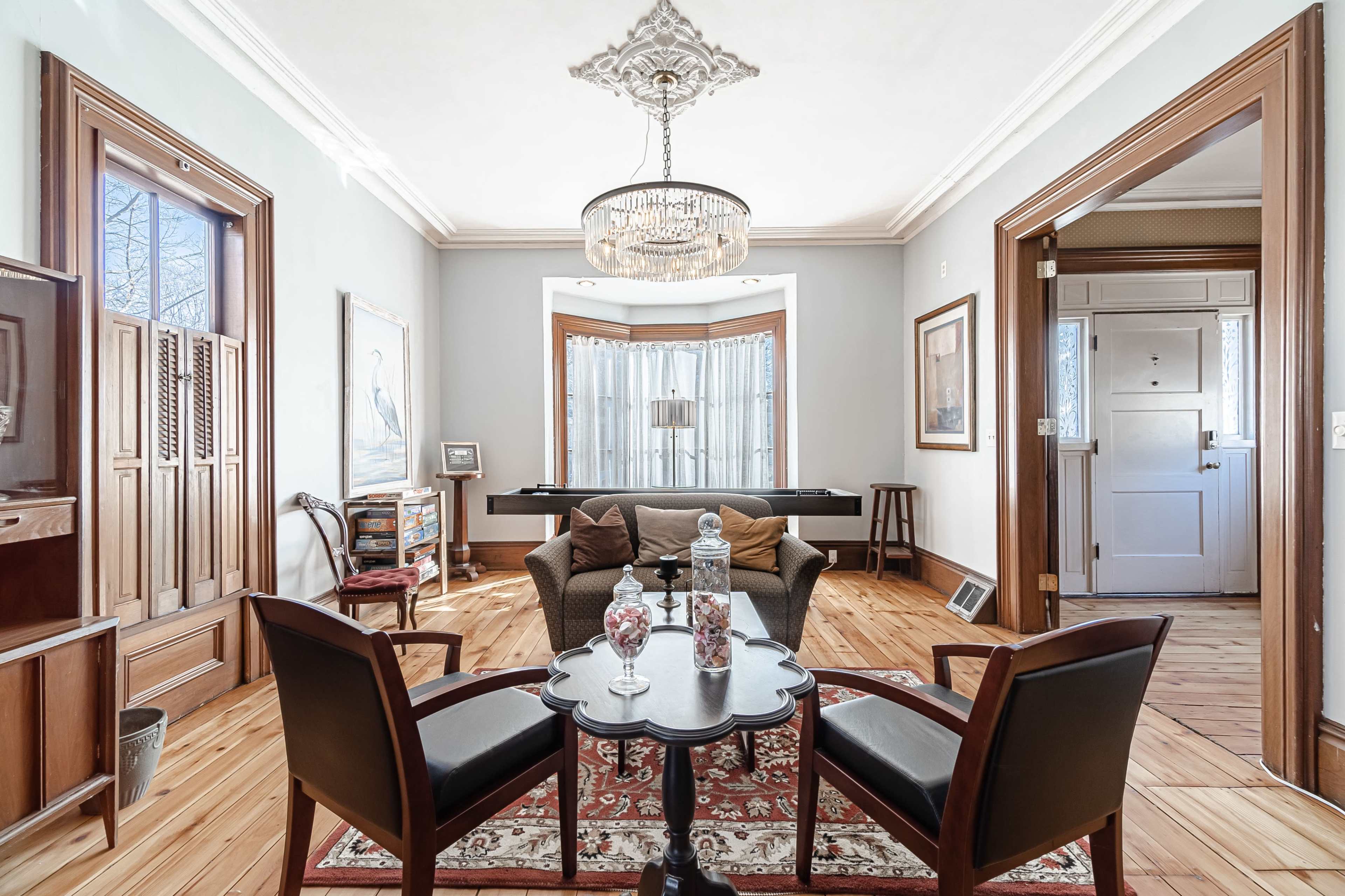 A well-furnished sitting area with two chairs and a small table, featuring a decorative rug, a large window with drapes, and wooden accents.