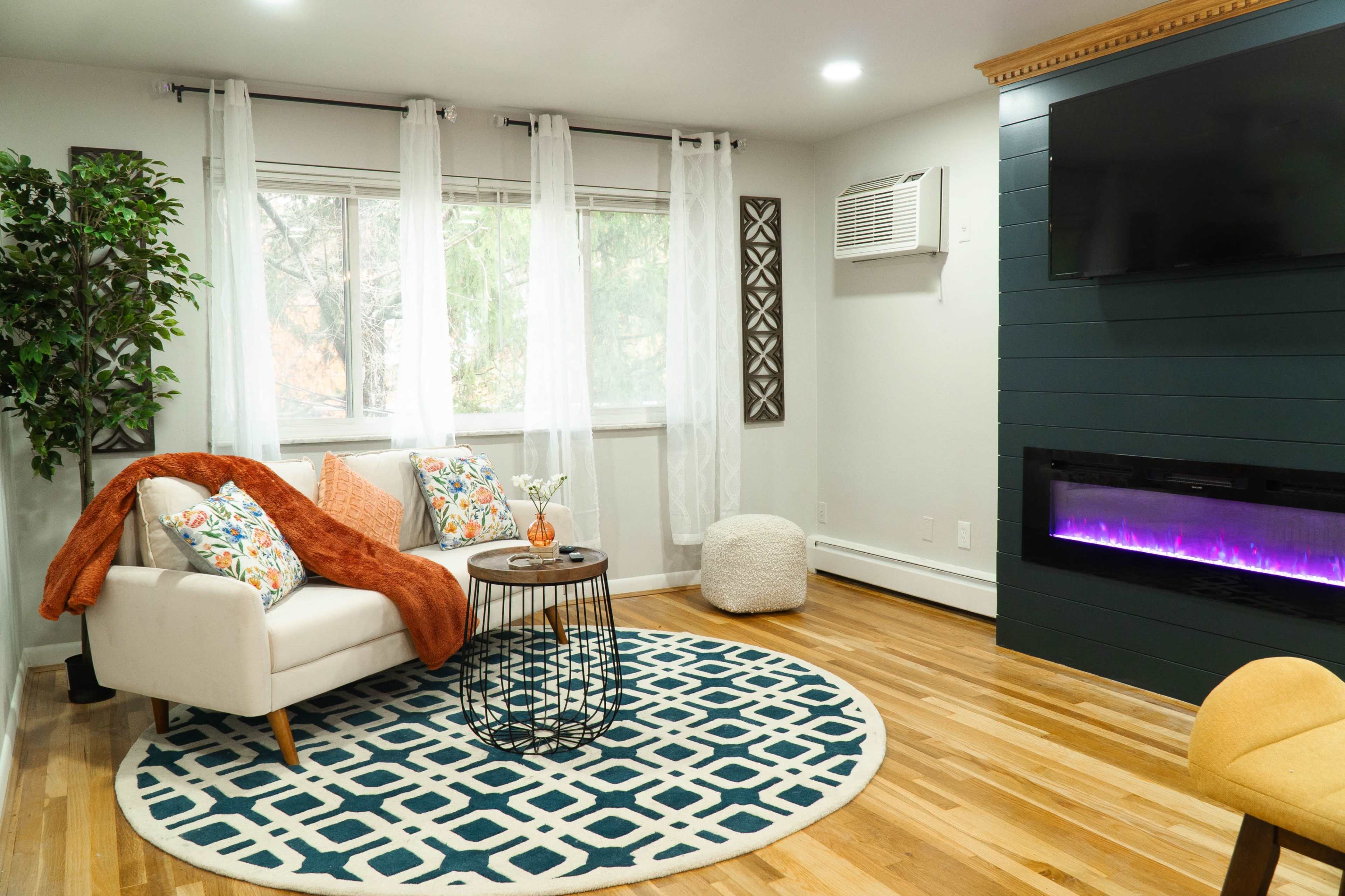 The living room features a white couch with colorful pillows, a round area rug, a small table, and a modern electric fireplace with a dark feature wall.