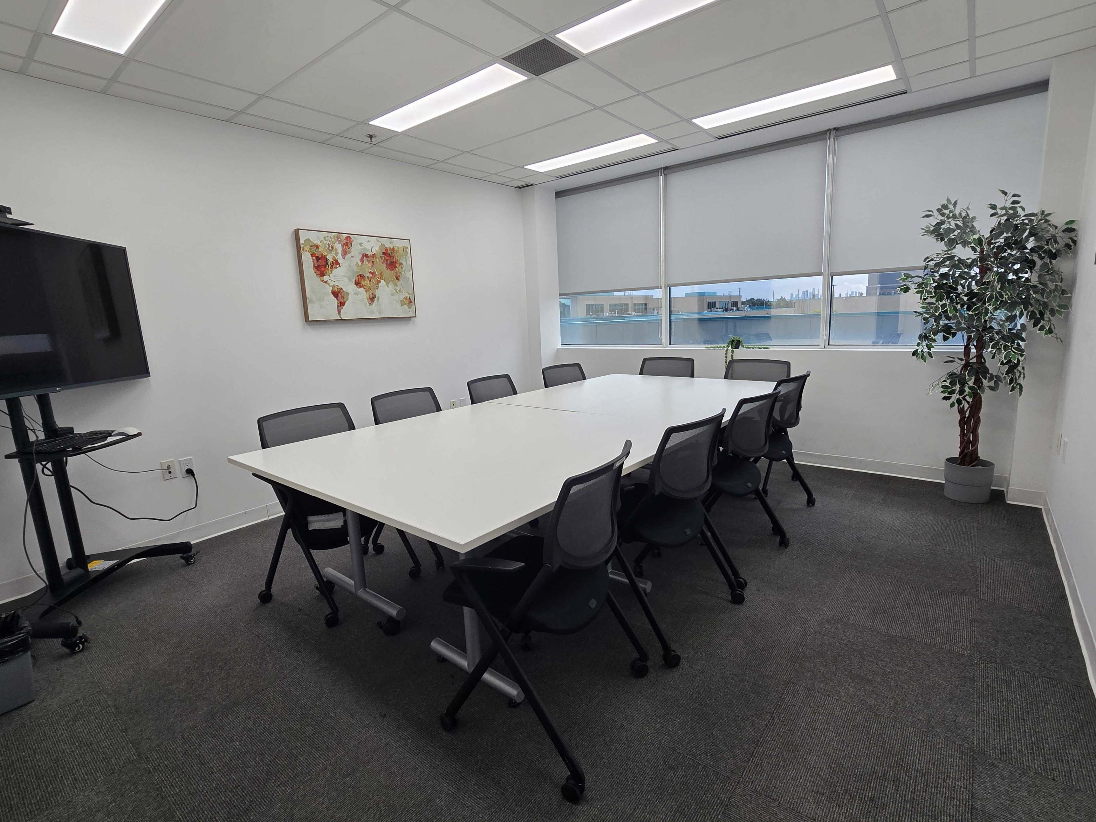 A modern conference room features a large white table surrounded by eight chairs, with a wall-mounted screen and a plant in one corner.