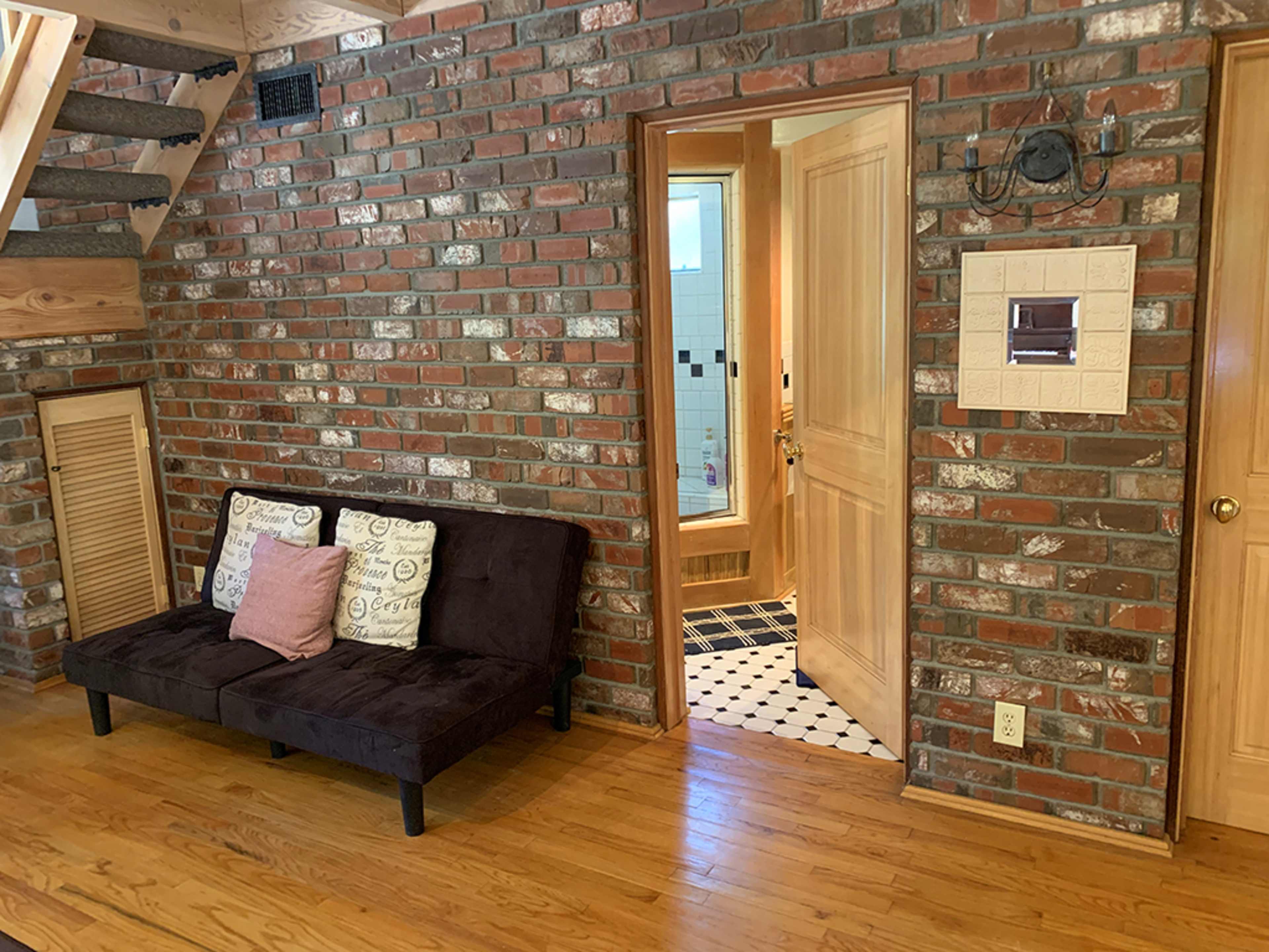 A room with a brick wall, a dark couch, and a doorway leading to a bathroom with tiled flooring.