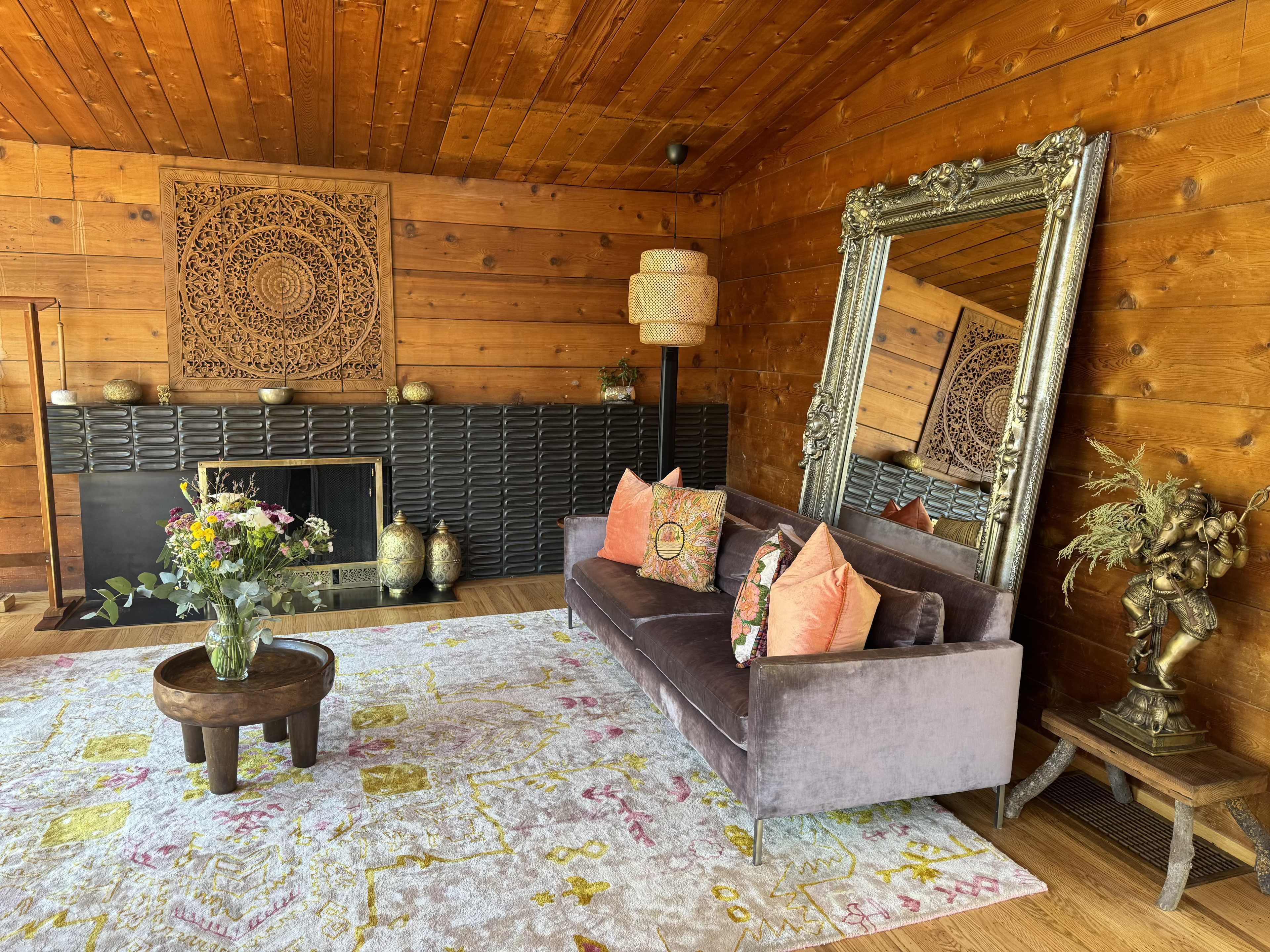 A cozy living room with a wooden ceiling, a velvet sofa with decorative pillows, a large mirror, and a floral rug, accompanied by various decorative elements on a fireplace and side table.