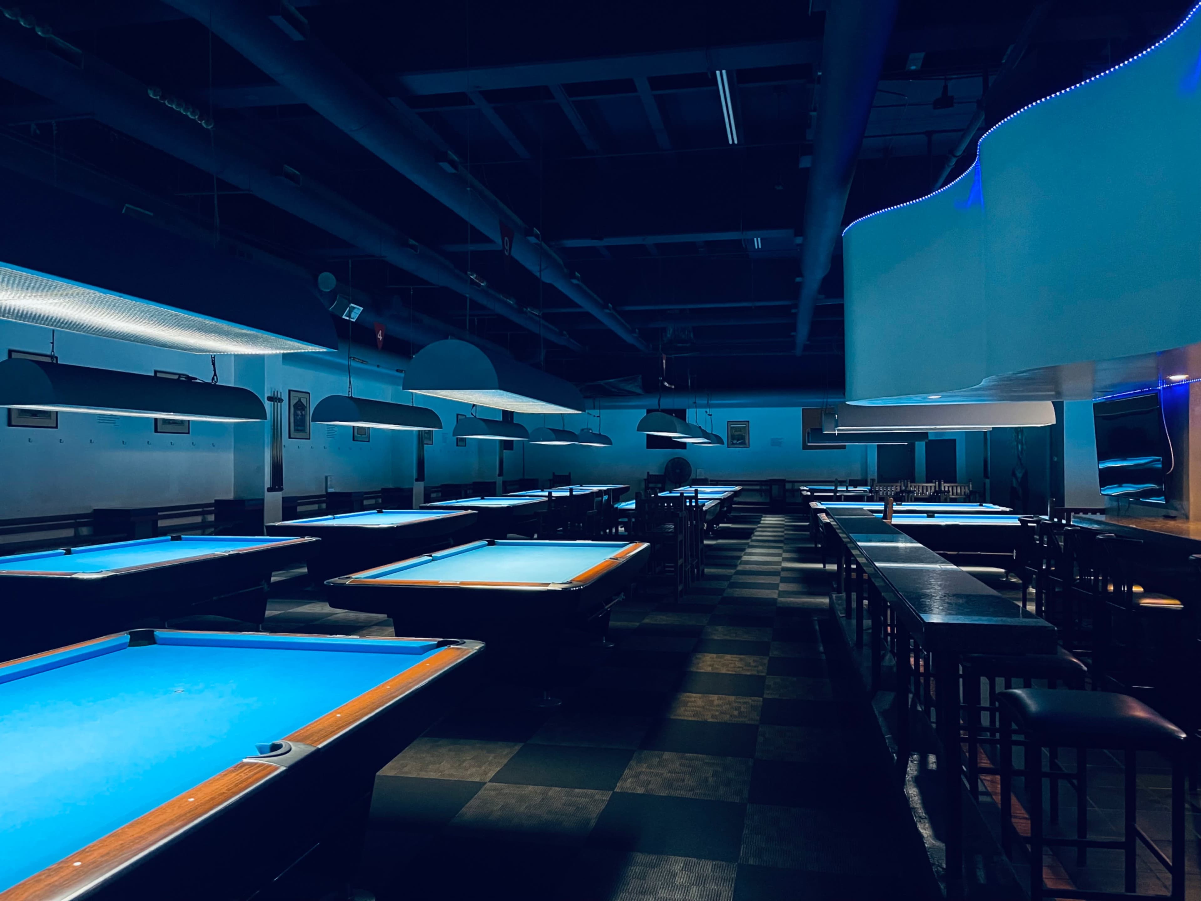 A dimly lit pool hall features several tables with blue felt and empty seating areas.