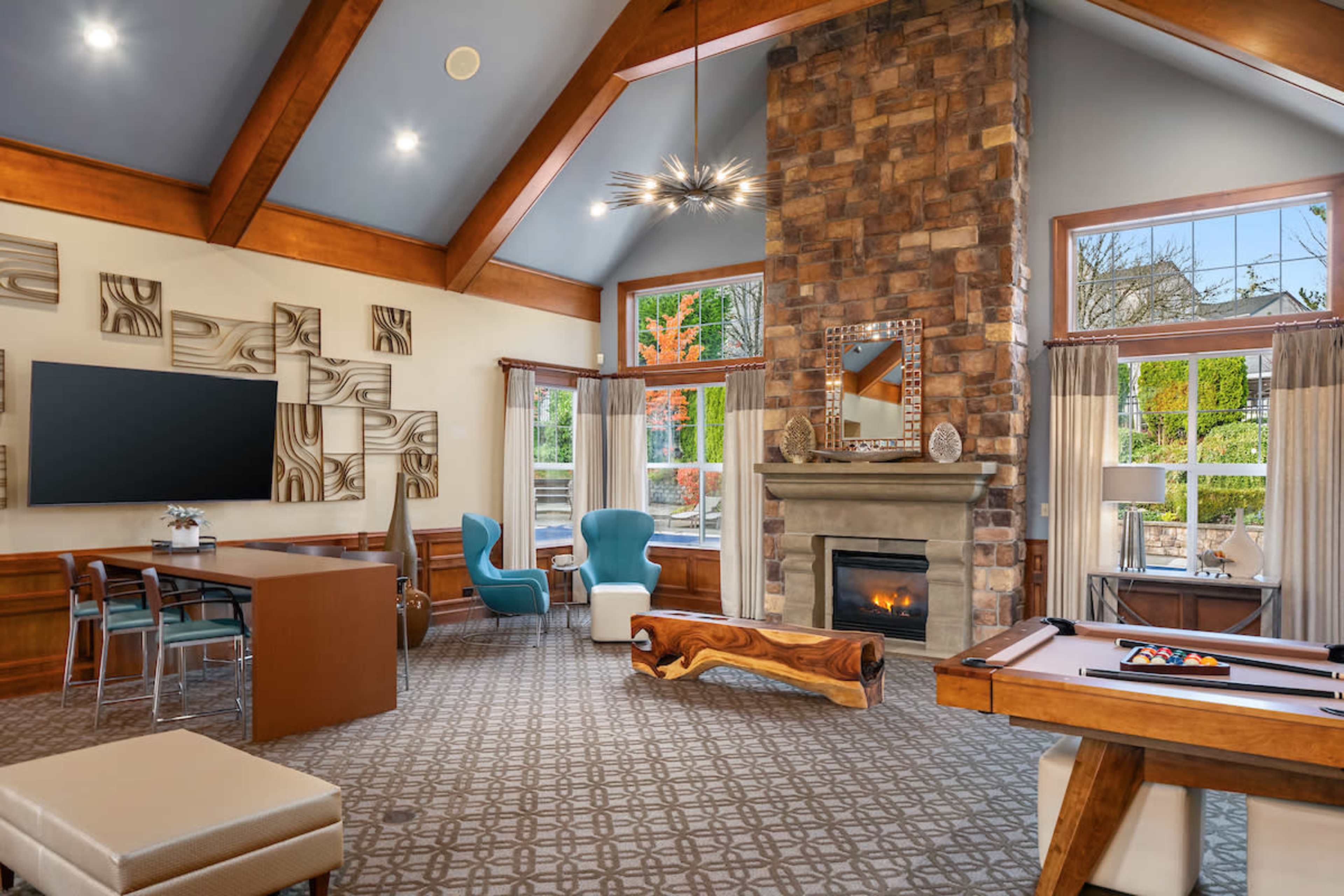 The image shows a spacious lounge area featuring a stone fireplace, large windows, modern furniture, and a game table.