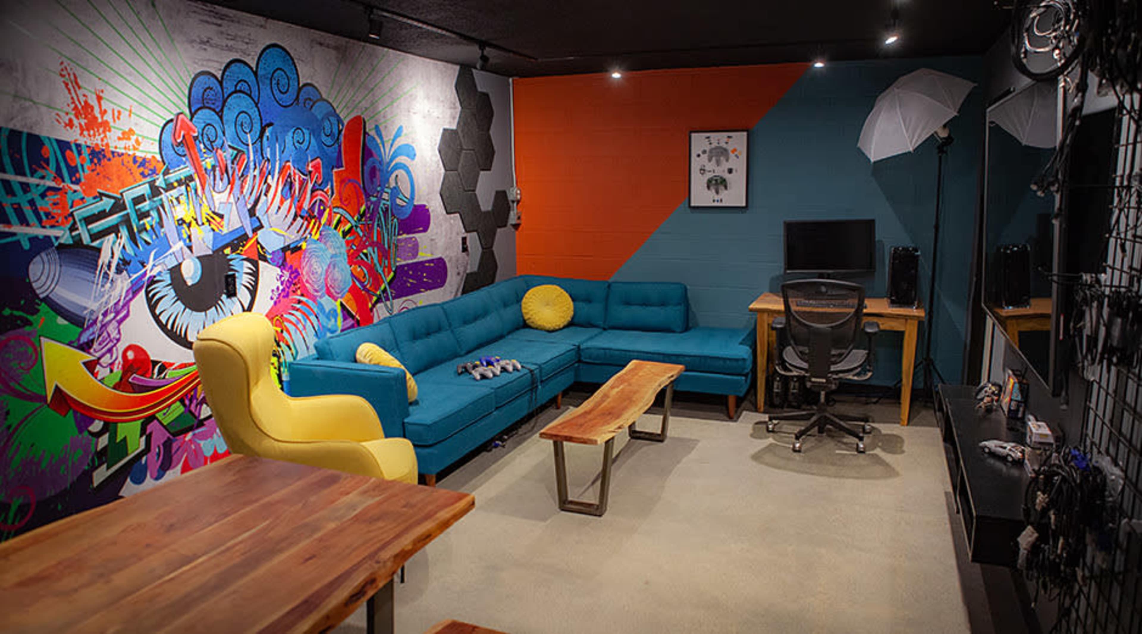 The image shows a modern lounge space featuring a colorful mural, a blue sectional sofa, a wooden coffee table, a desk with a computer, and soft lighting.