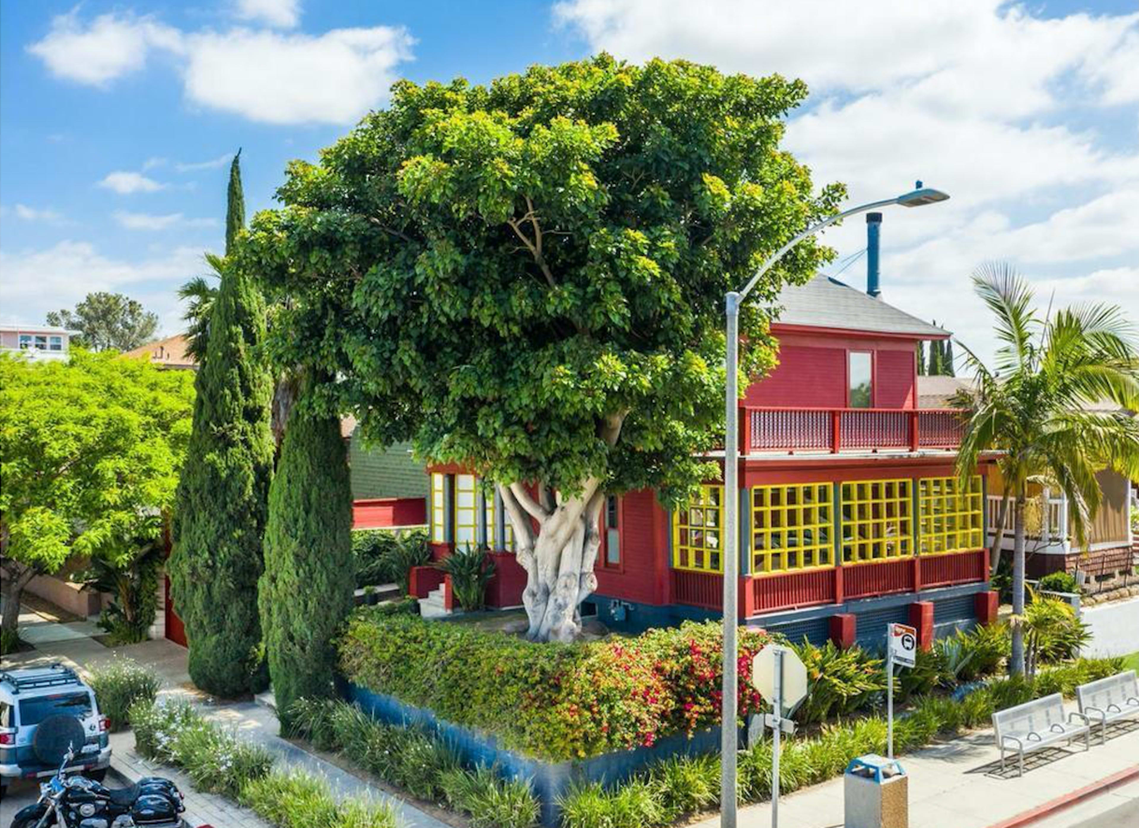 A large red house with yellow-trimmed windows is situated on a street, surrounded by various trees and greenery.