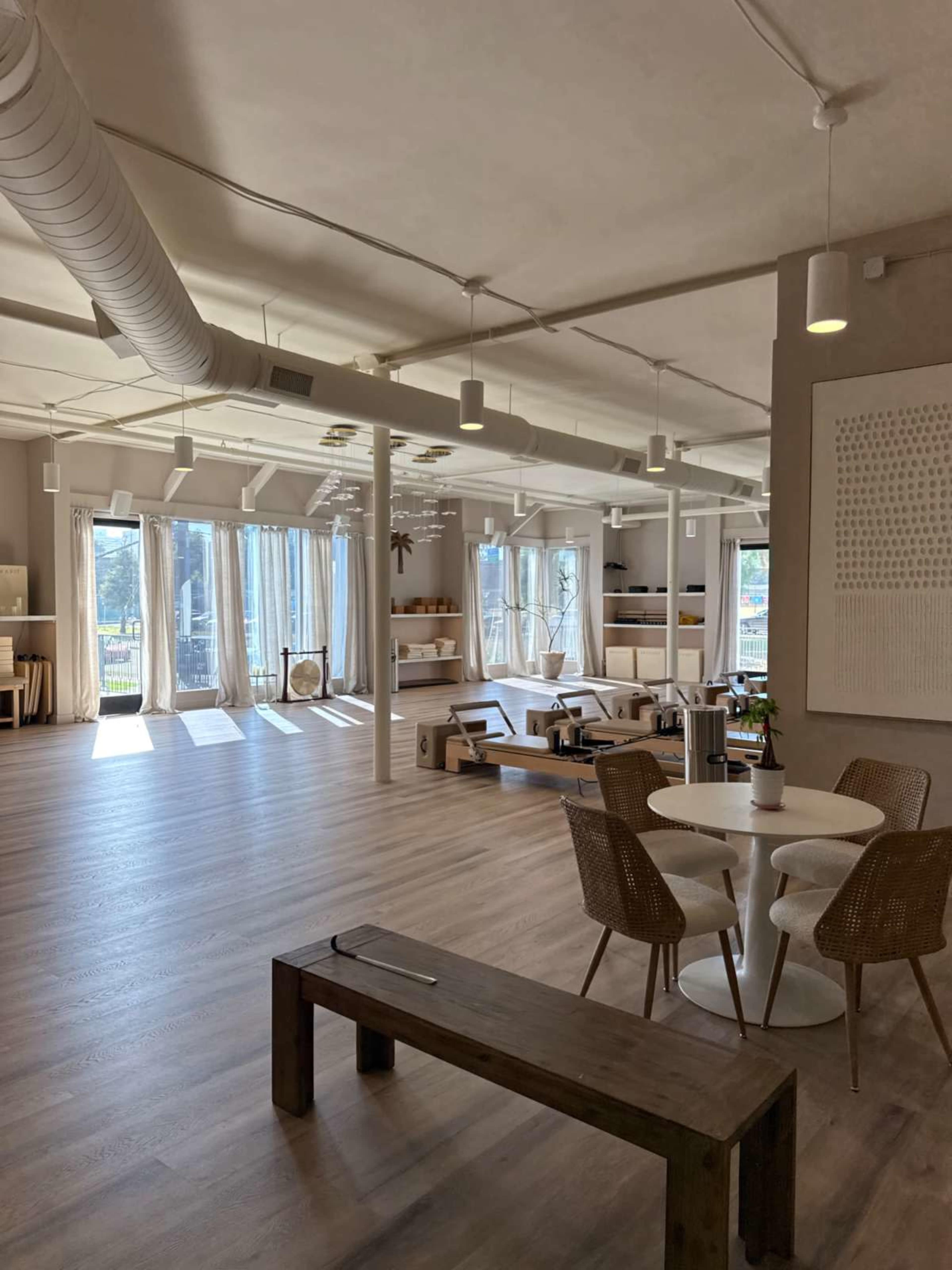 The image shows a well-lit, modern interior space with large windows, wooden furniture, and a minimalist design featuring a seating area and dining tables.