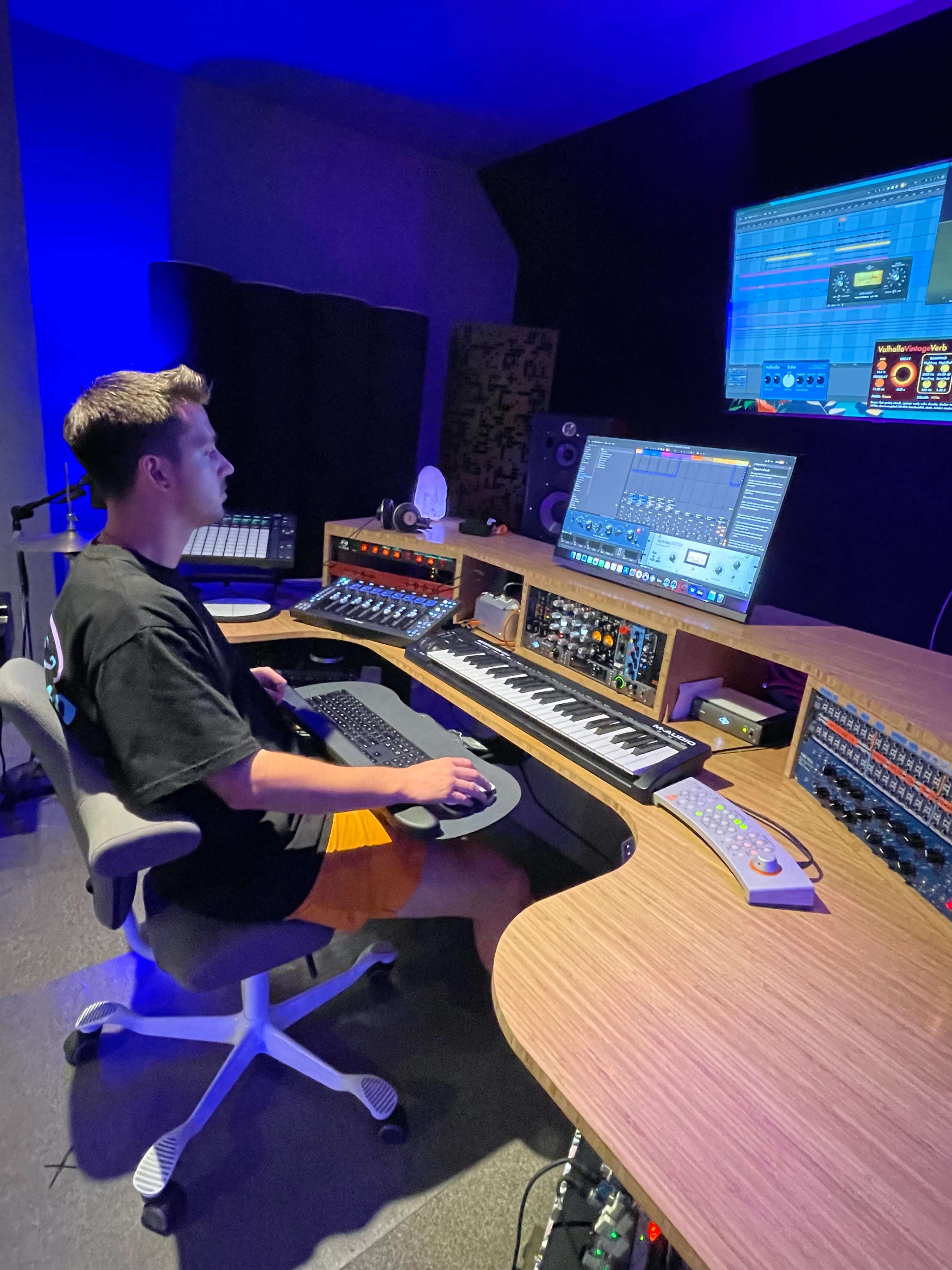 A person sits at a music production workstation, using a computer and mixing console in a dimly lit studio.