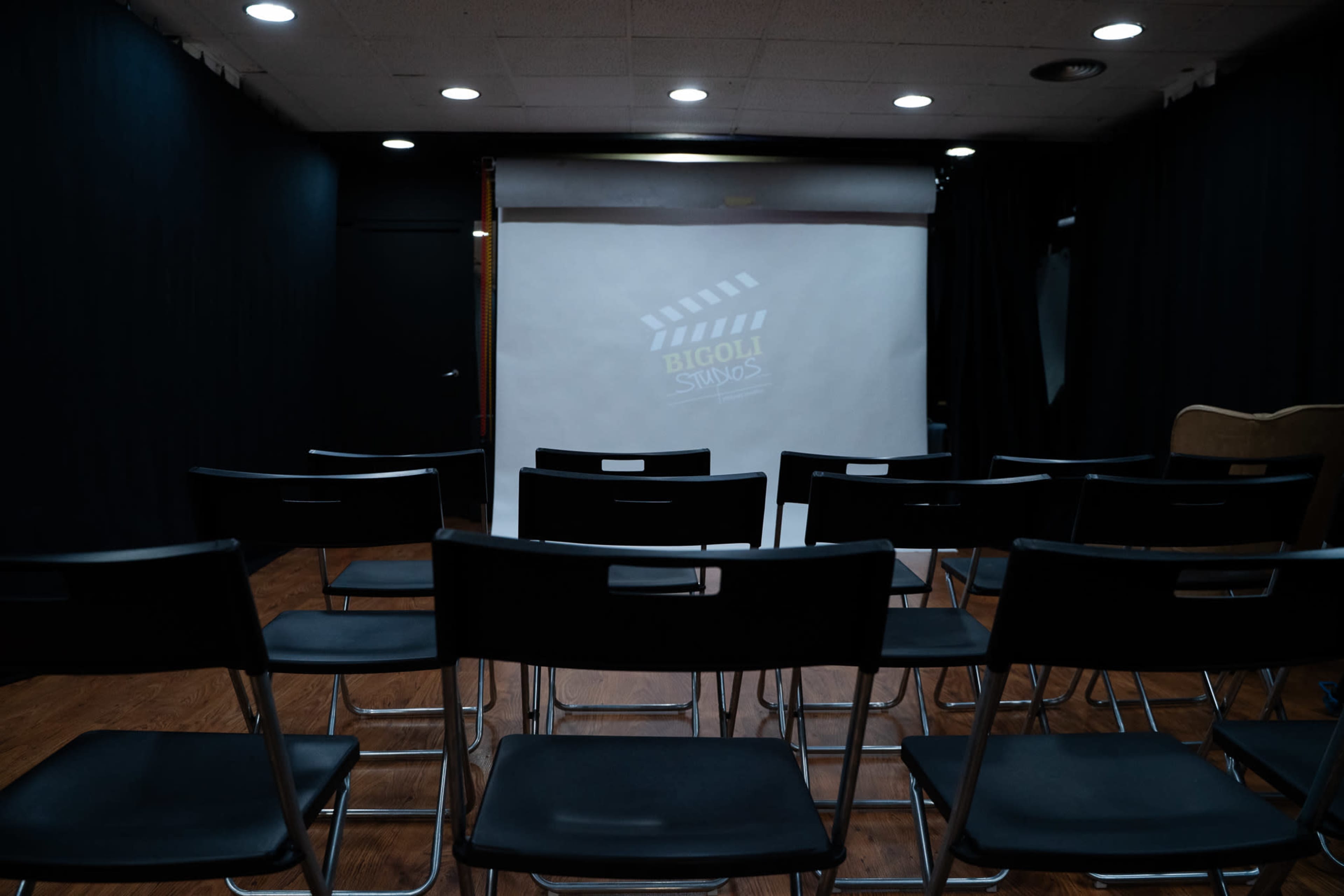 The scene shows a small room with a projector screen displaying a film clapperboard logo and several rows of empty black folding chairs facing the screen.