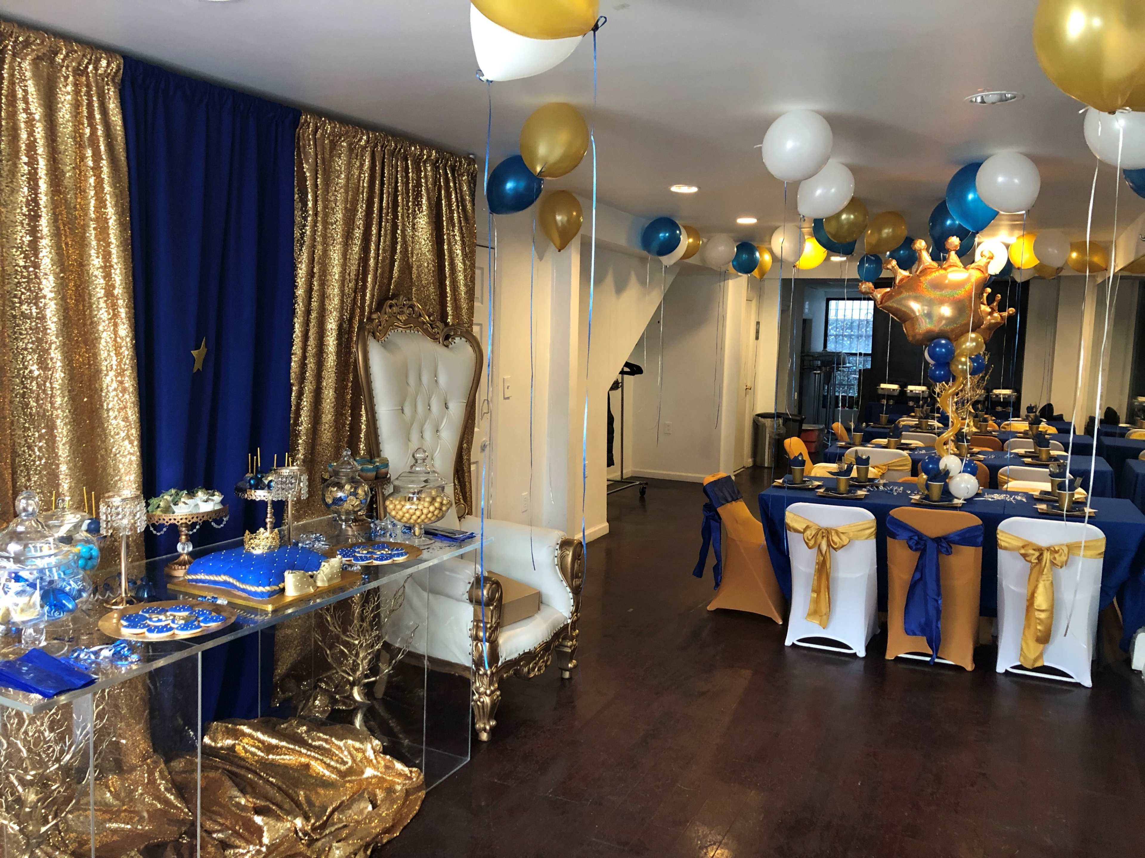 A decorated event space featuring a dessert table with golden drapery, white and blue balloons, and elegantly set tables with blue and gold tablecloths.