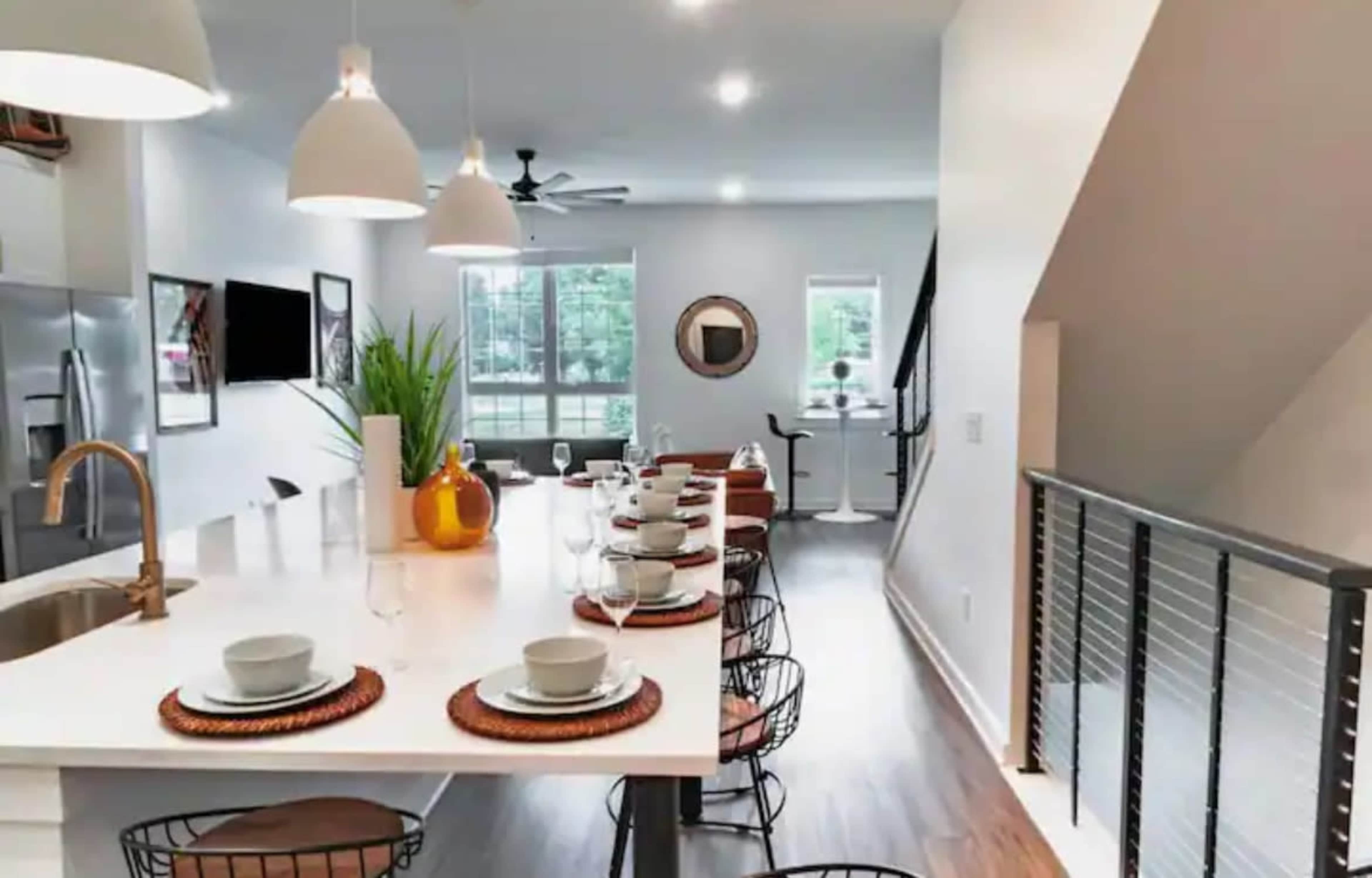 A modern kitchen and dining area features a long white countertop with table settings and a staircase leading to the upper level.