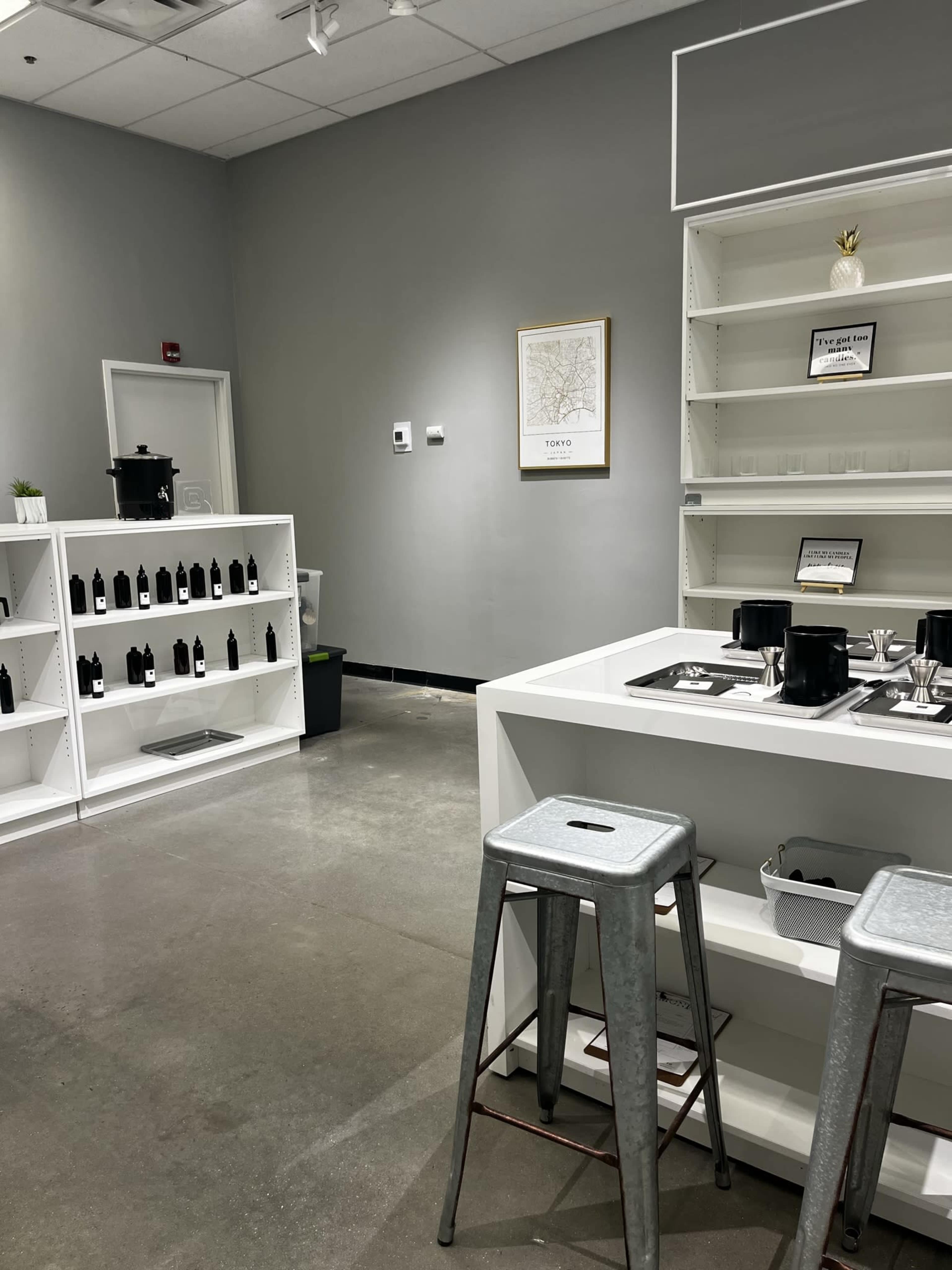 A minimalist indoor space with white shelves containing black bottles, a light-colored wall featuring a framed map, and a central white table with metal stools.