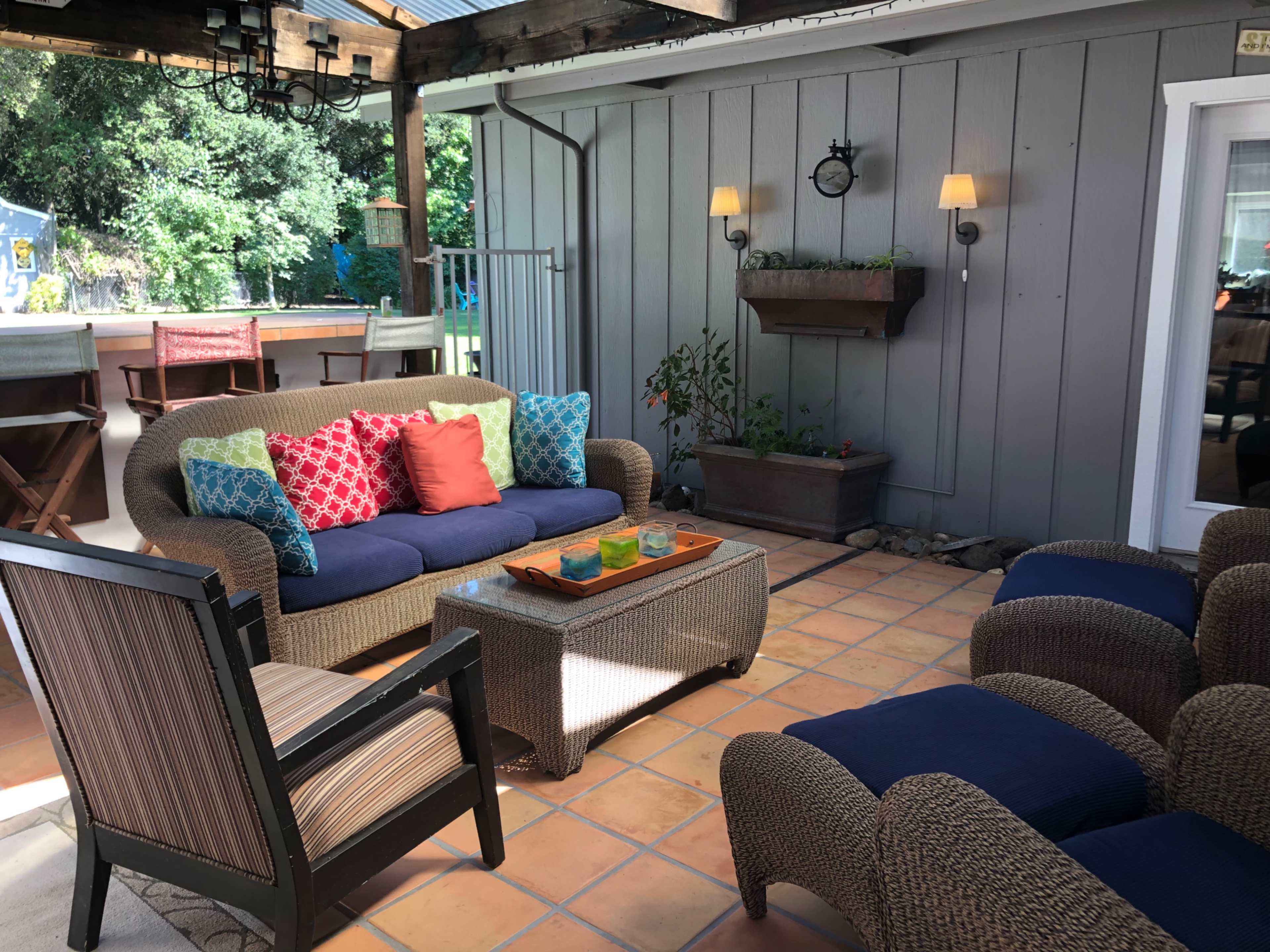 The scene features a patio area with a woven sofa adorned with colorful cushions, a wooden coffee table with decorative items, and surrounding chairs on a tiled floor.