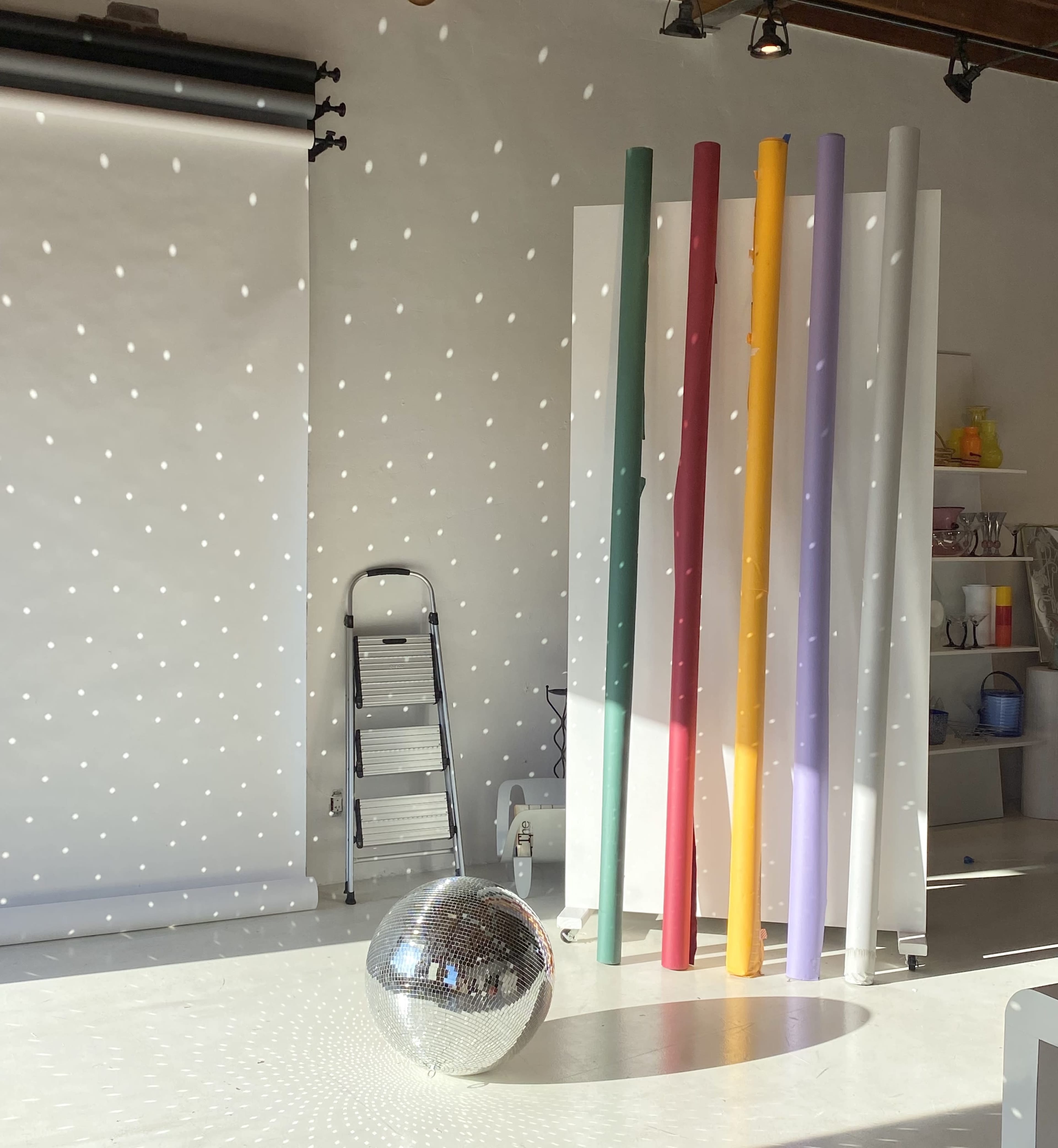 The scene features a bright studio with a mirrored disco ball on the floor and colorful rolls of paper leaning against a white backdrop.