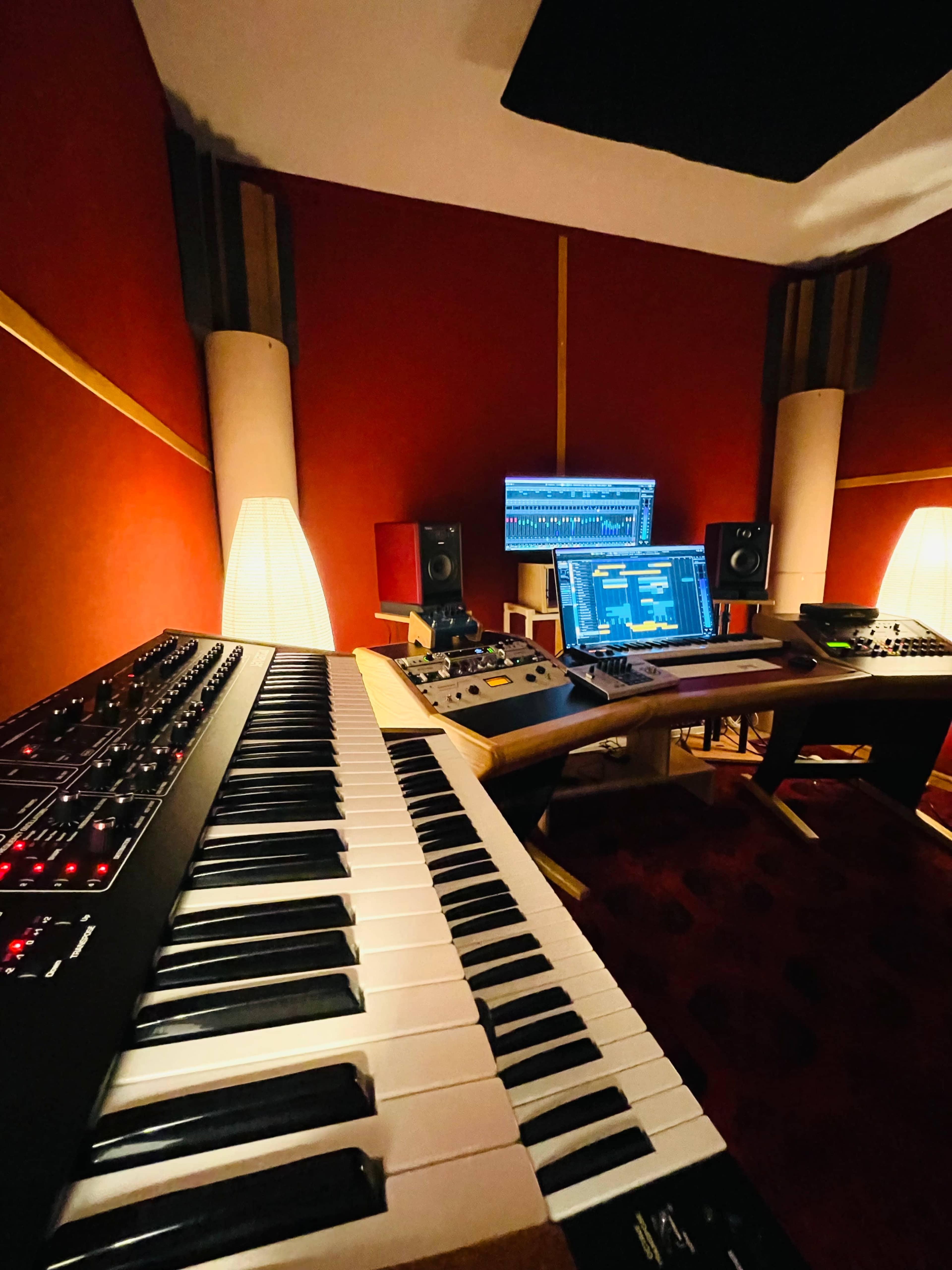 A music production studio with a keyboard, computer monitor, and audio equipment against a red wall.