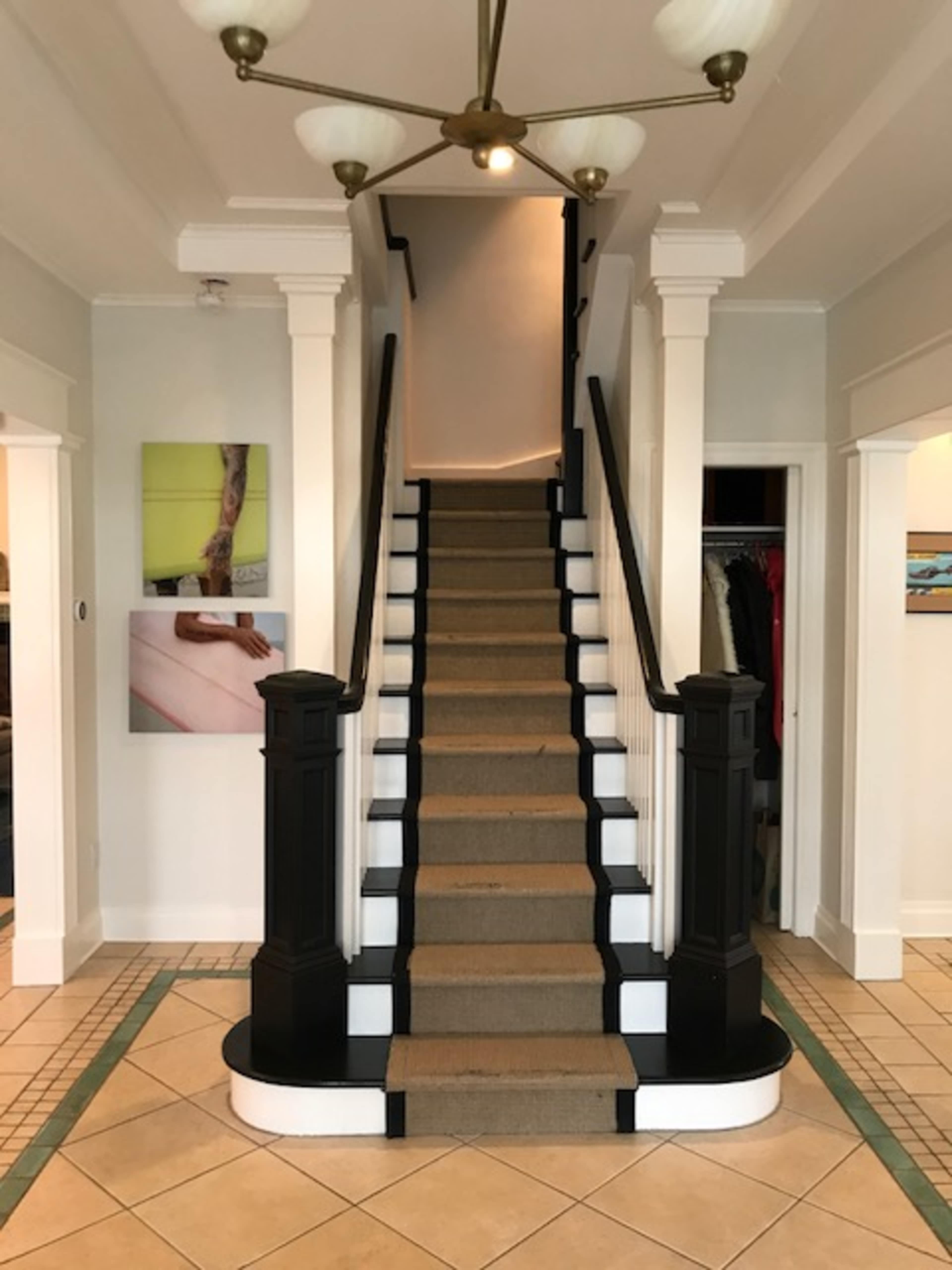 The image shows an indoor staircase with a black and white railing, leading upward, flanked by walls featuring artwork and a closet to the side.
