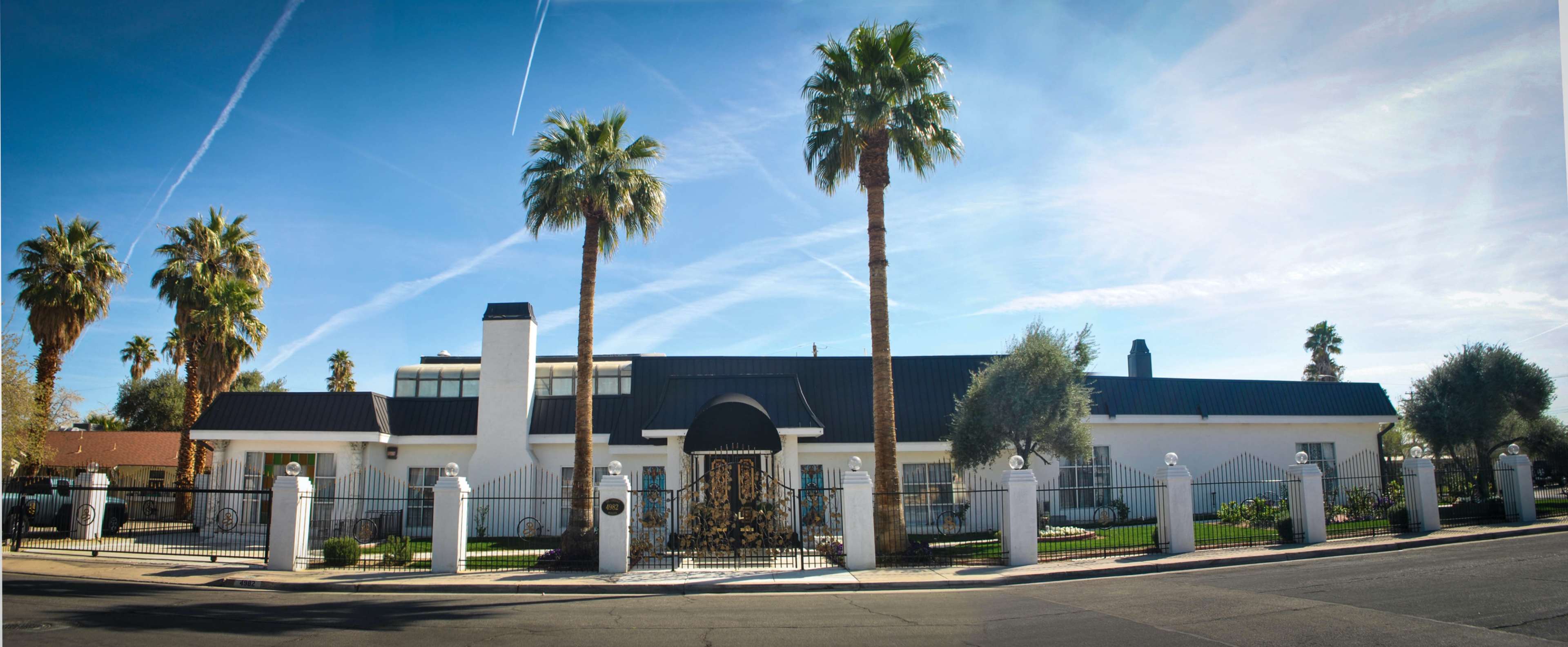 A modern white house with a black roof is surrounded by palm trees, featuring a gated entrance and well-maintained landscaping.