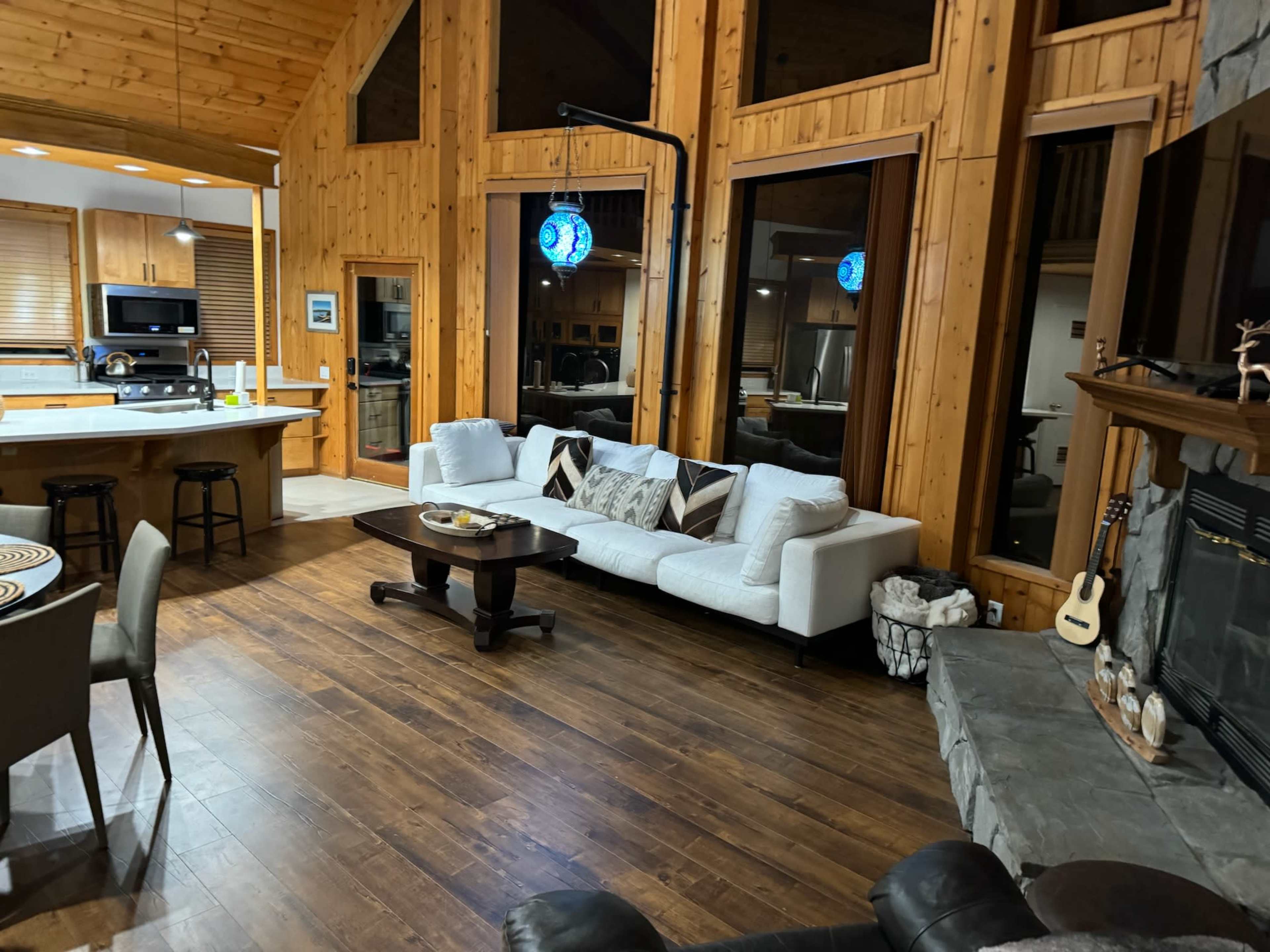 The image shows a warm, wooden interior of a living space featuring a white couch, a coffee table, and a stone fireplace, with a kitchen area visible in the background.