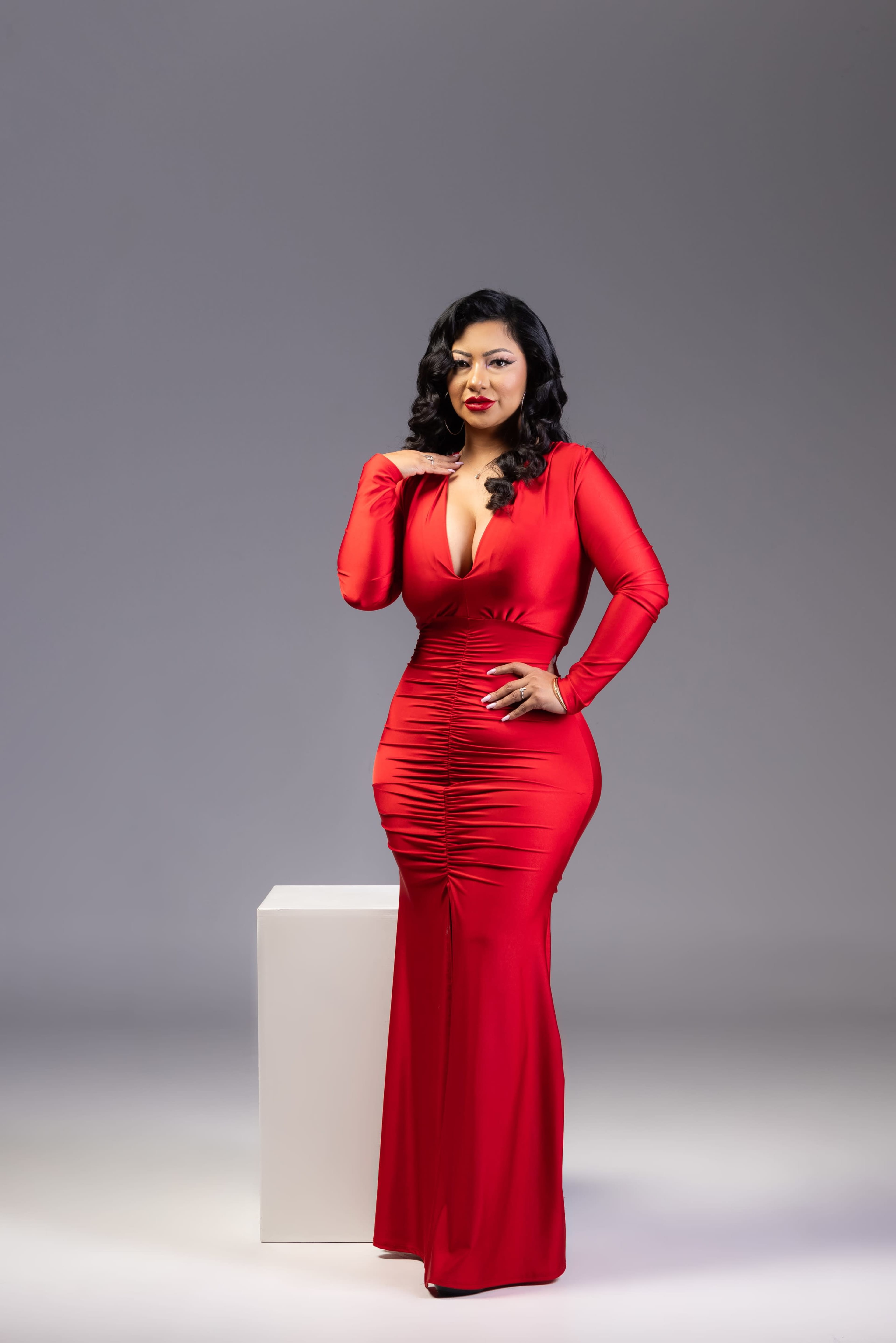 A woman poses confidently in a fitted red dress while standing beside a white pedestal.