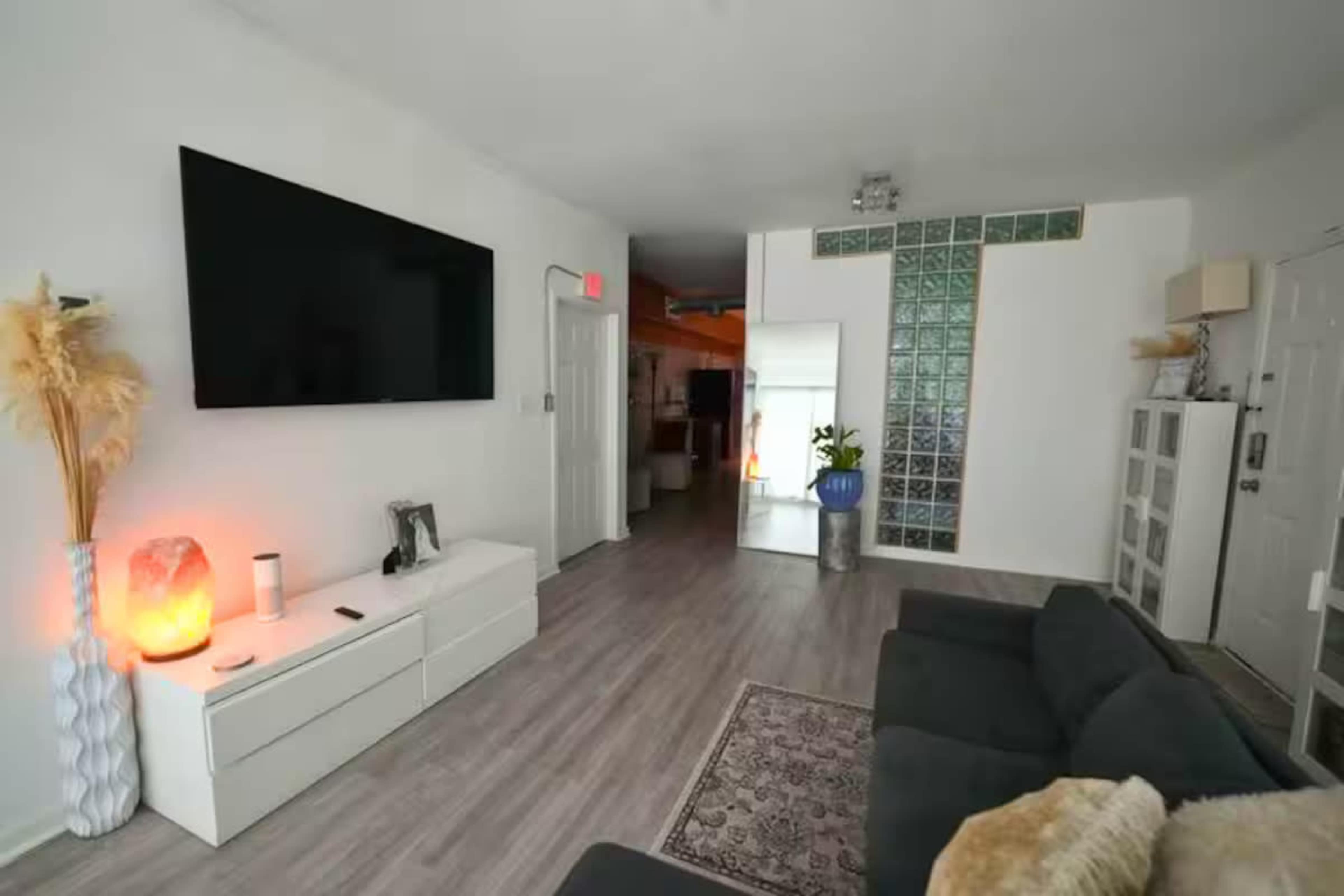 A modern living room features a large flat-screen TV mounted on the wall, a white media console, a dark sofa, and a decorative glass block wall.