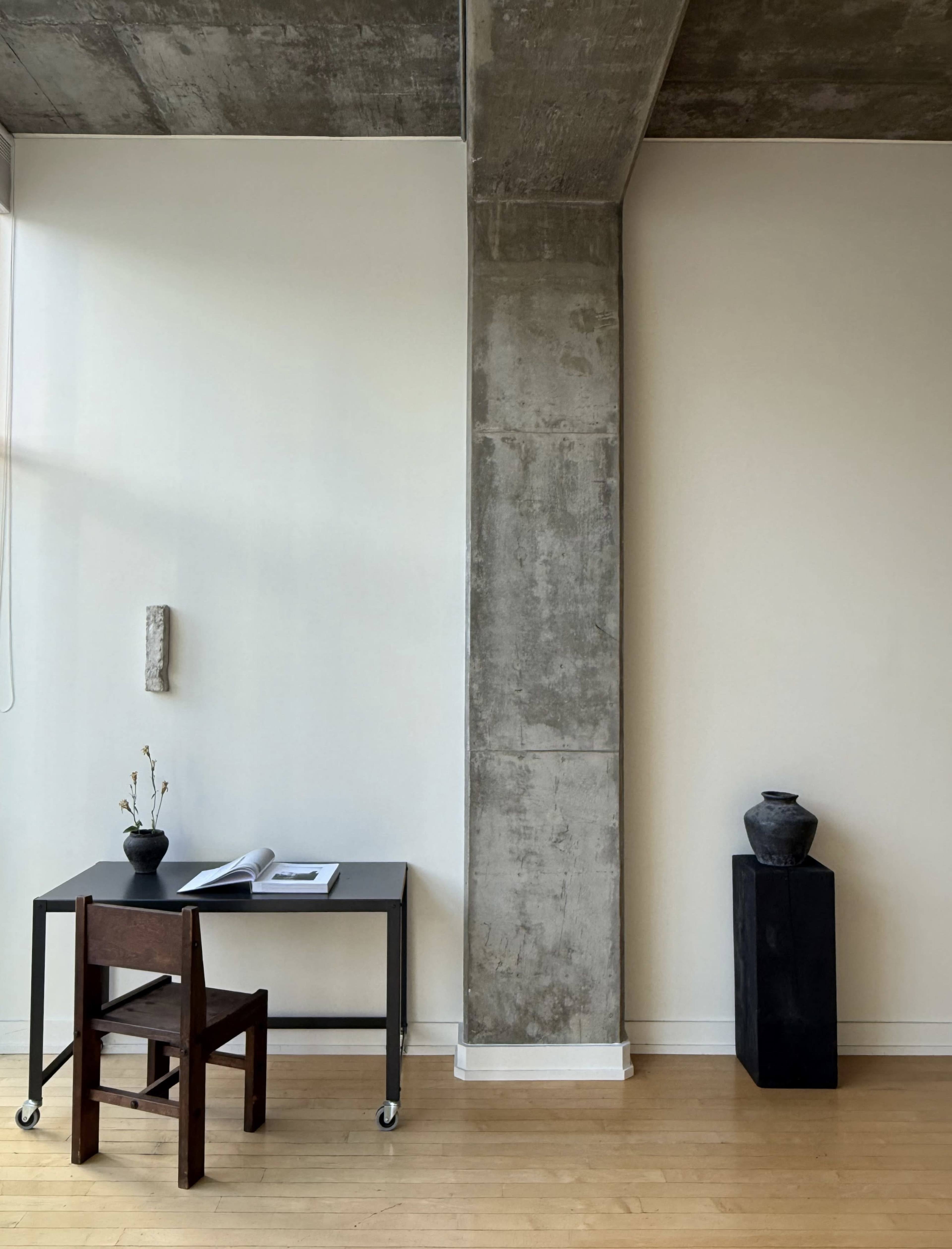A minimalist room features a wooden chair and a black table next to a concrete pillar, with a small vase and a sculpture on display.