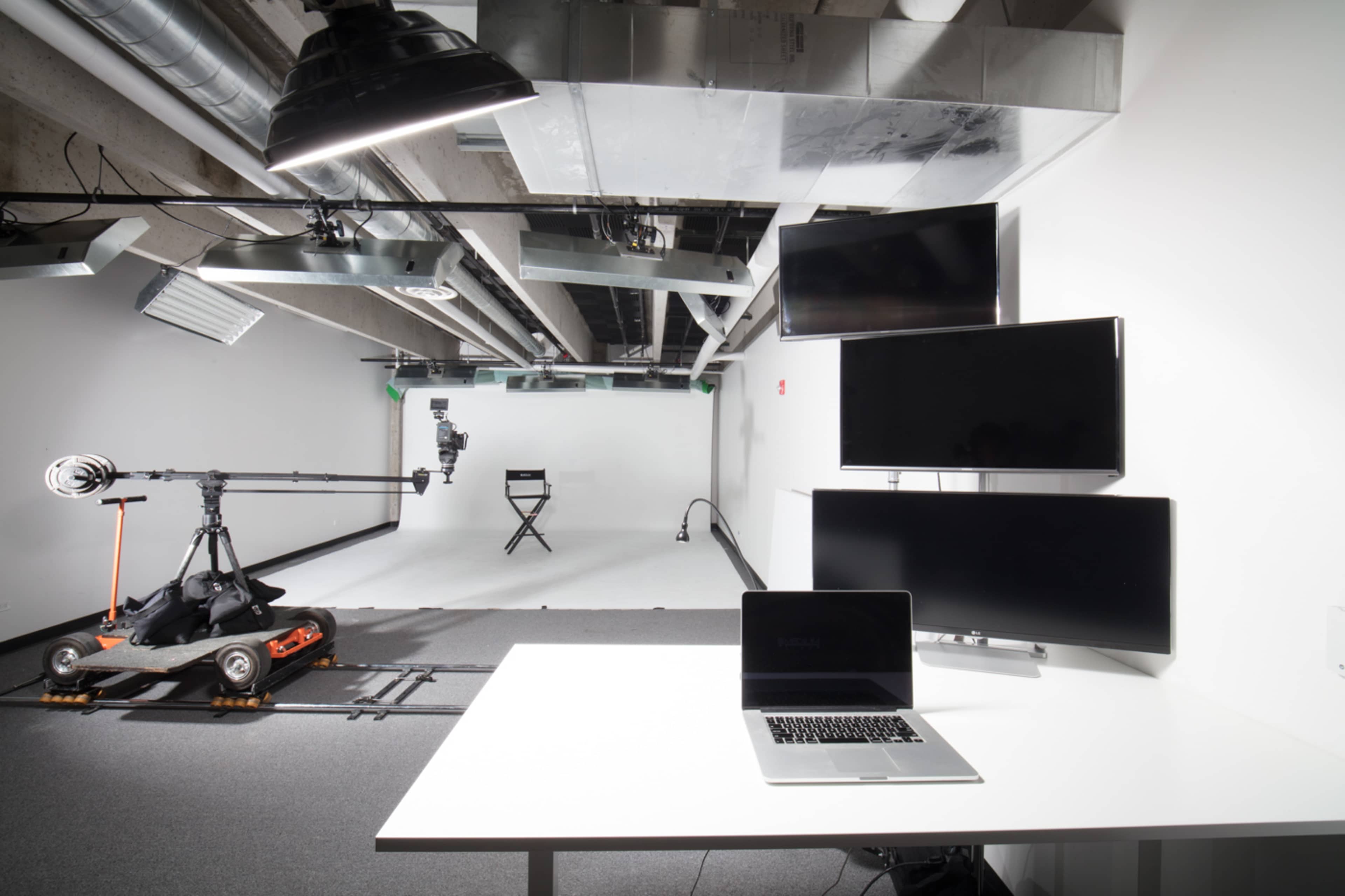 A modern studio setup featuring a white backdrop, a camera mounted on a dolly, multiple monitors, and a laptop on a white table.