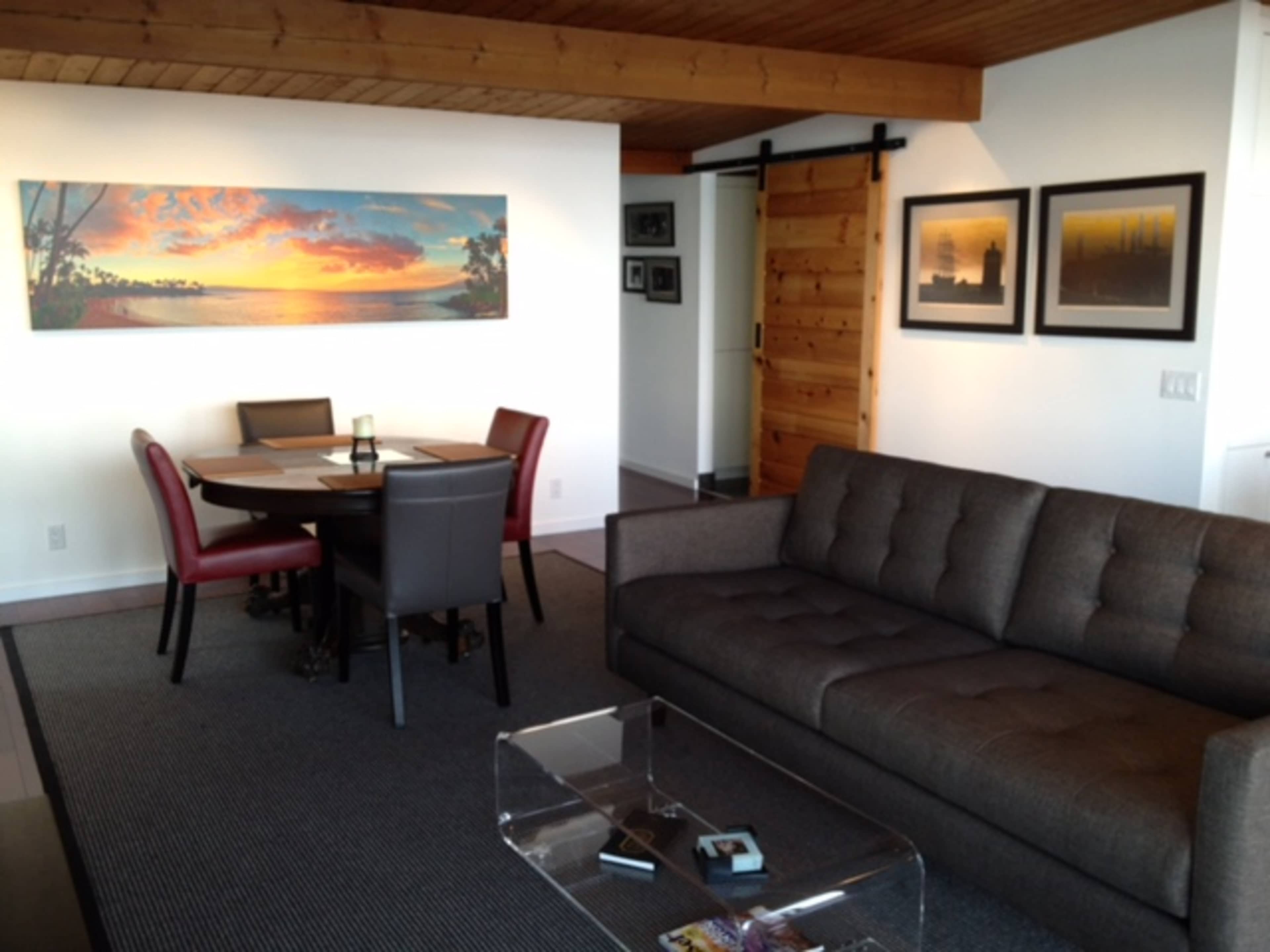 A modern living space with a gray sofa, a glass coffee table, a dining area with a circular table and chairs, and a large wall art piece depicting a sunset.