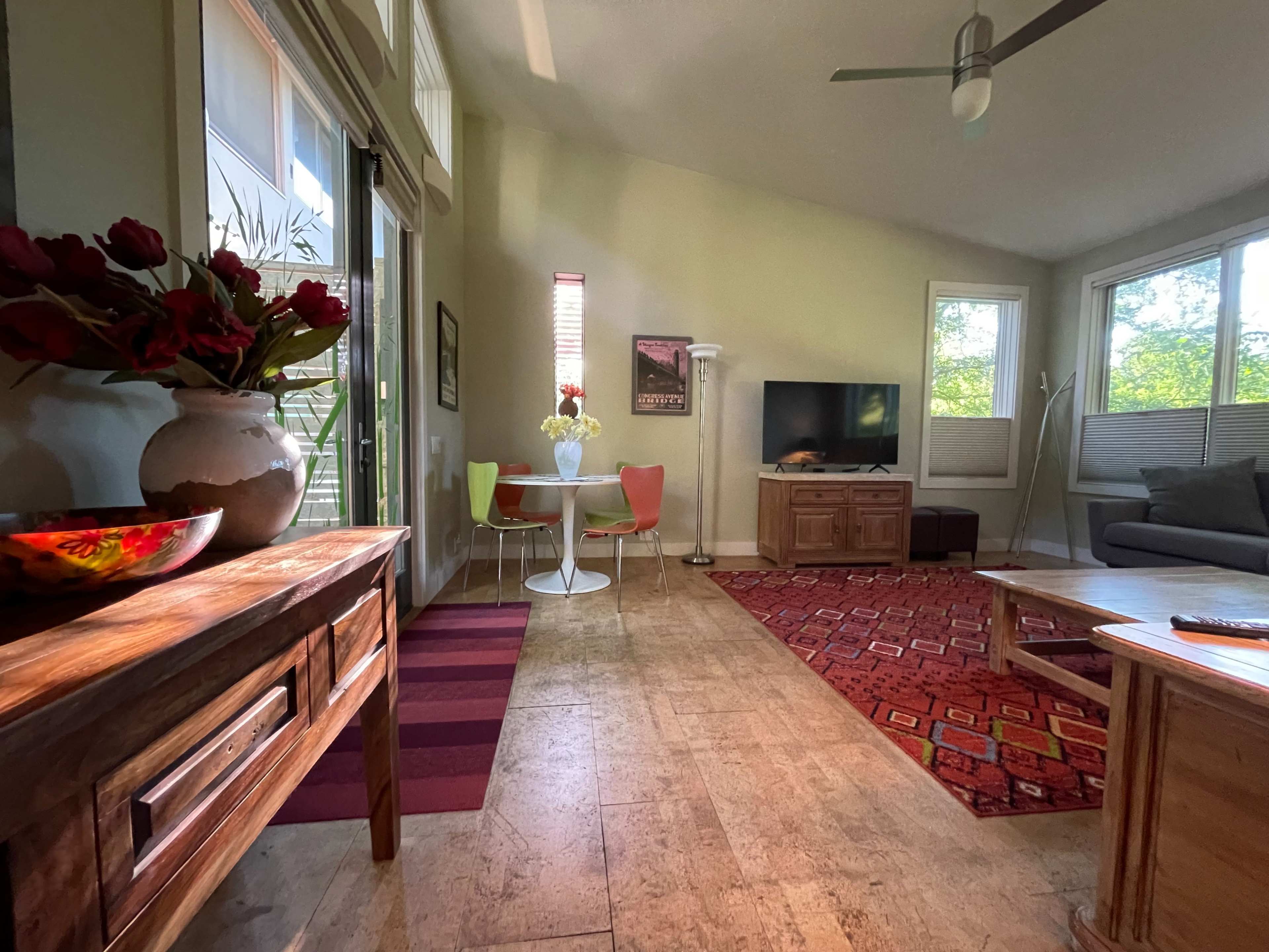 A bright living space features a wooden sideboard, a dining area with colorful chairs, a TV, and large windows allowing natural light.