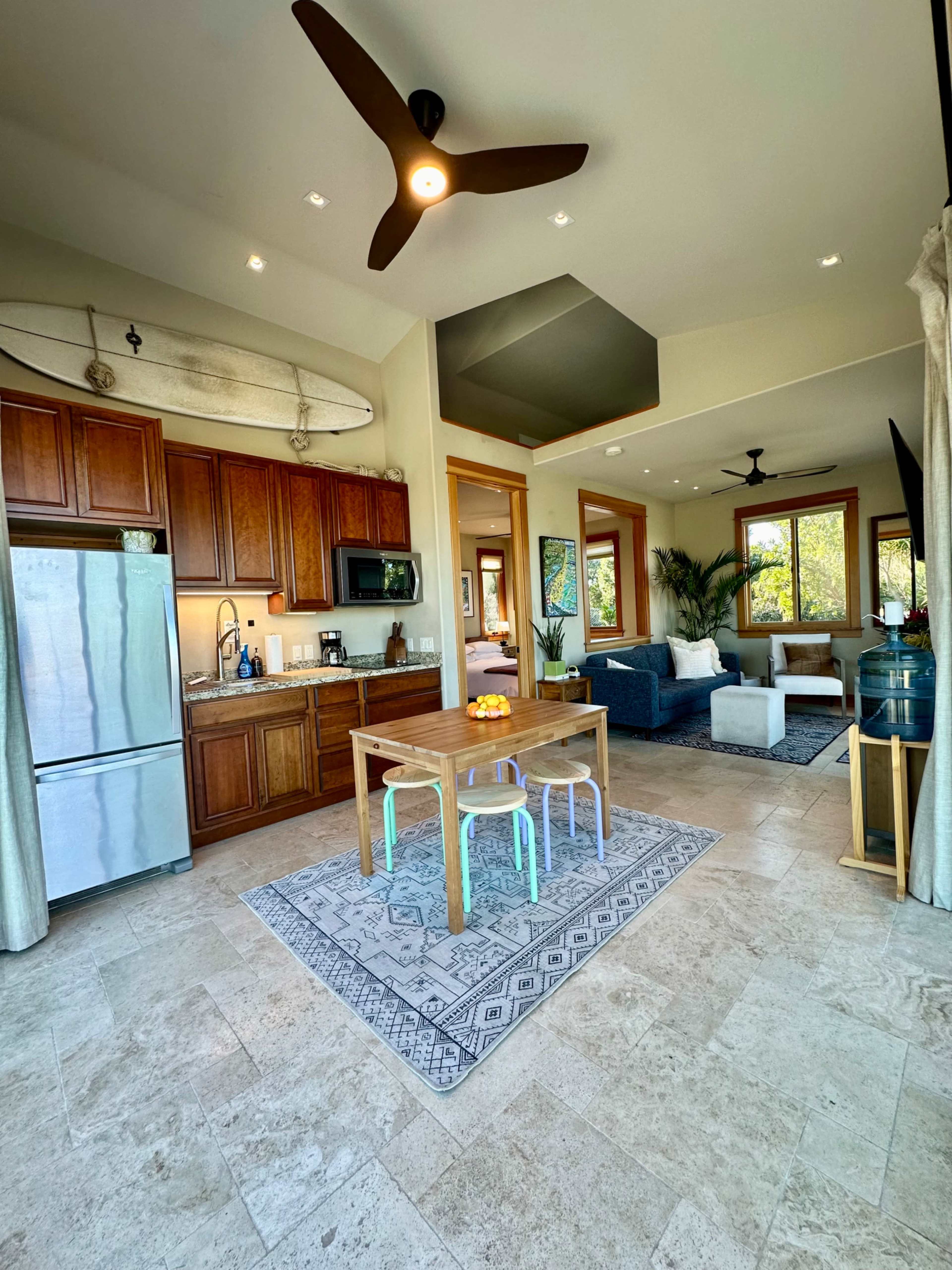 The image shows a modern open-concept living space featuring a kitchen with wooden cabinets, a dining table with colorful chairs, and a cozy seating area with large windows.