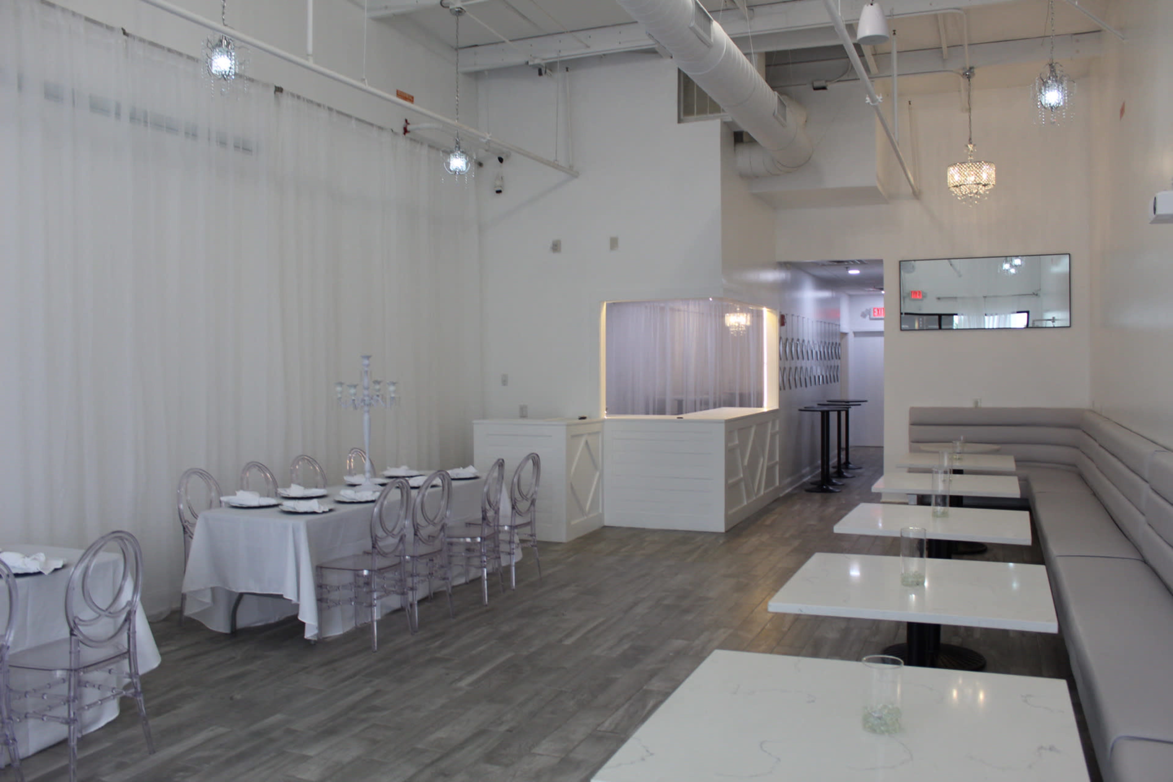 The image shows a minimalist restaurant interior featuring white walls, light wooden floors, and clear acrylic chairs arranged around white tables.