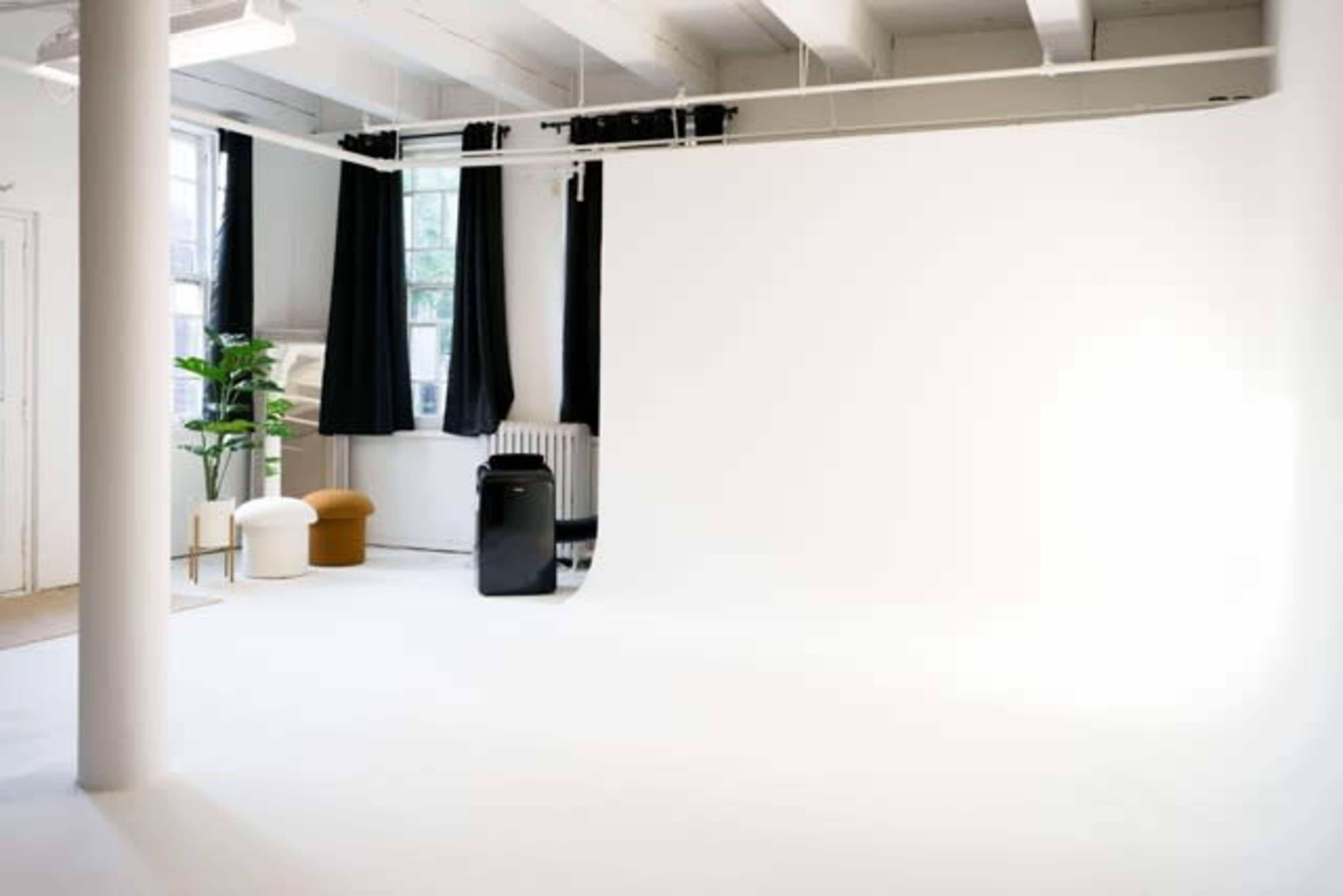 A spacious studio with a white backdrop, a green plant, and a few pieces of furniture under a bright ceiling.