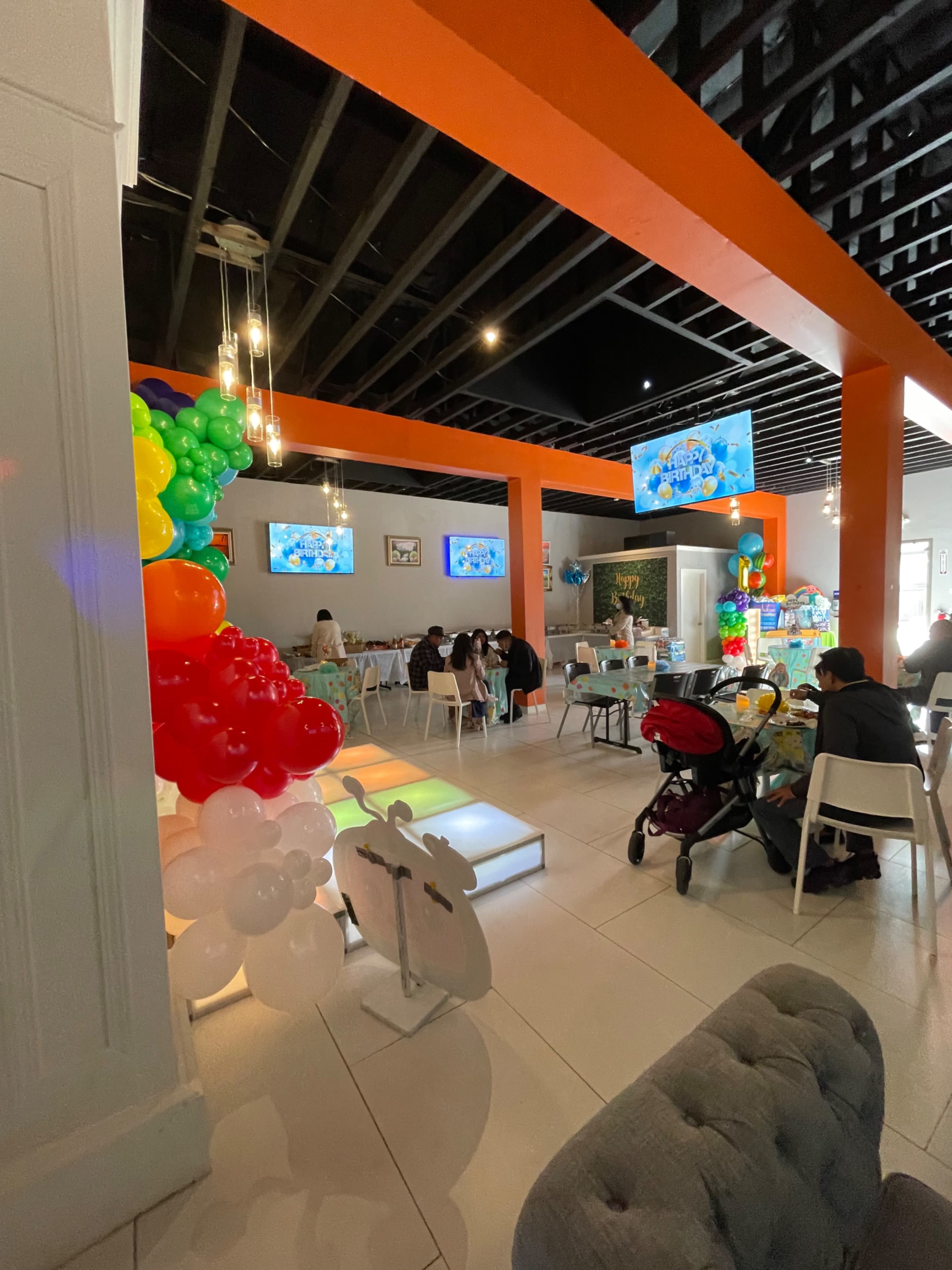 The image shows a brightly decorated indoor event space with colorful balloons, tables set for a celebration, and guests seated at various tables.
