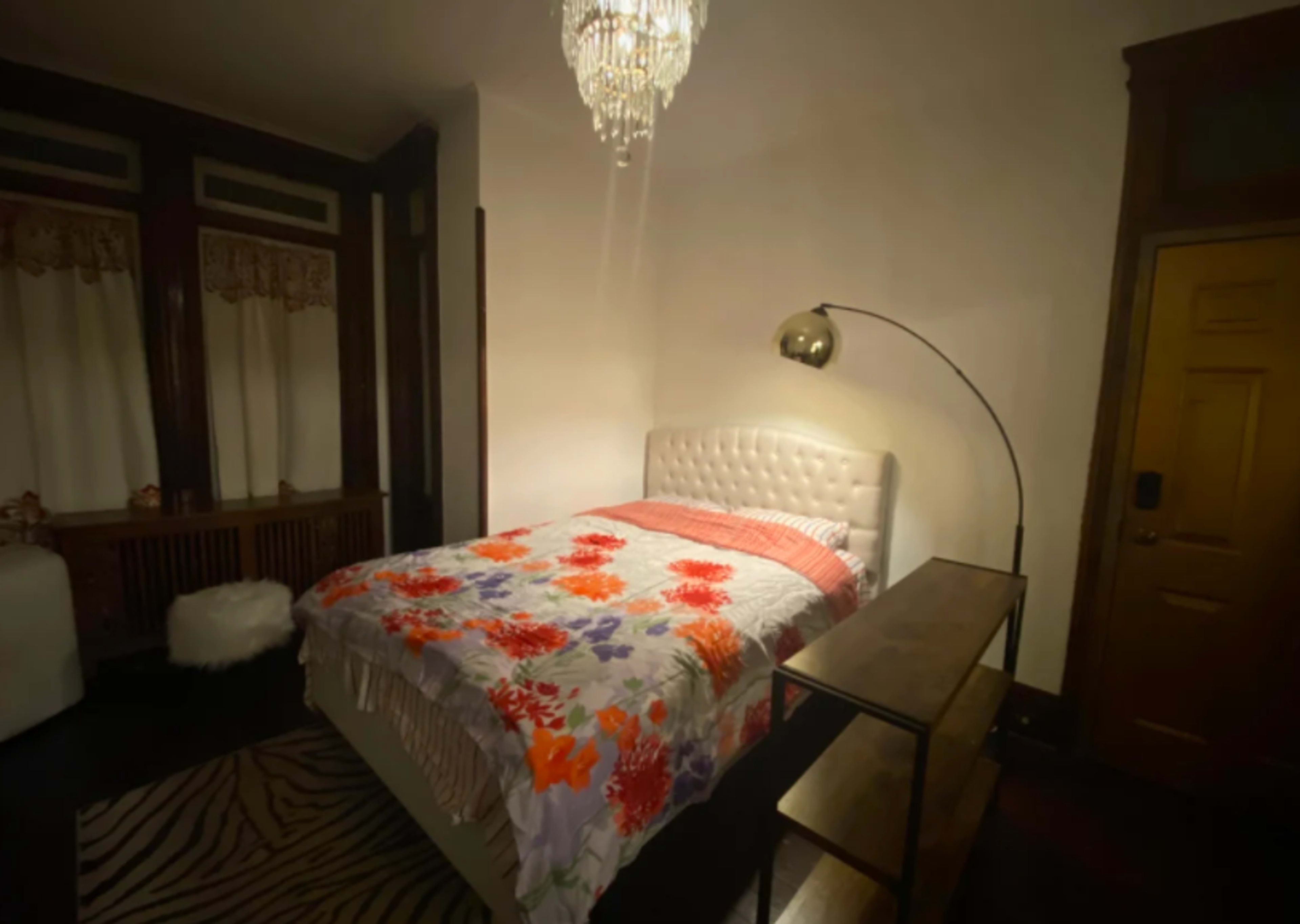 The bedroom features a tufted white bed with a floral quilt, an arc floor lamp, and wooden furnishings against a dark wall.