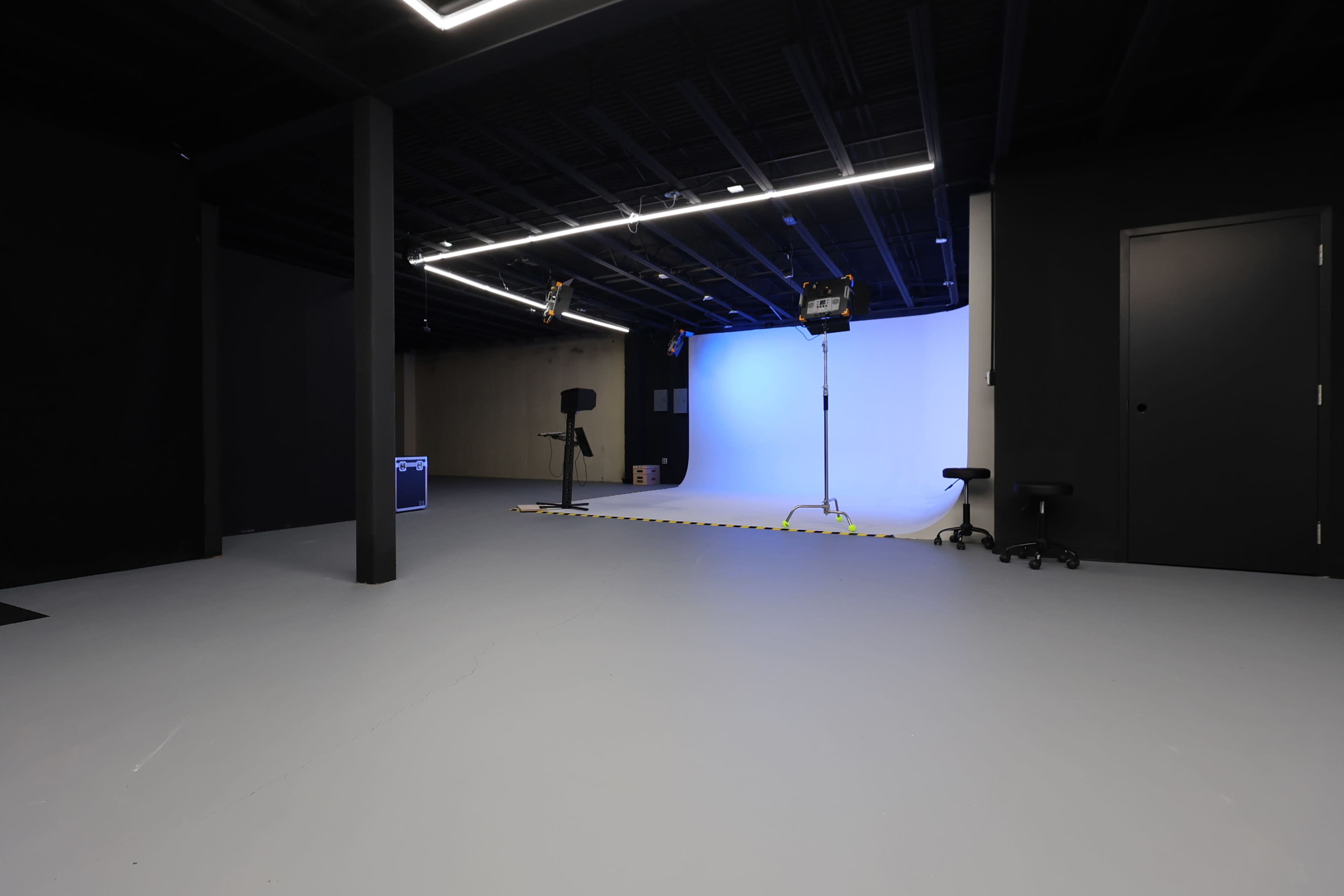 The image shows a spacious photo or video studio with a blue backdrop, a lighting setup, and minimal furniture, all under dim lighting.