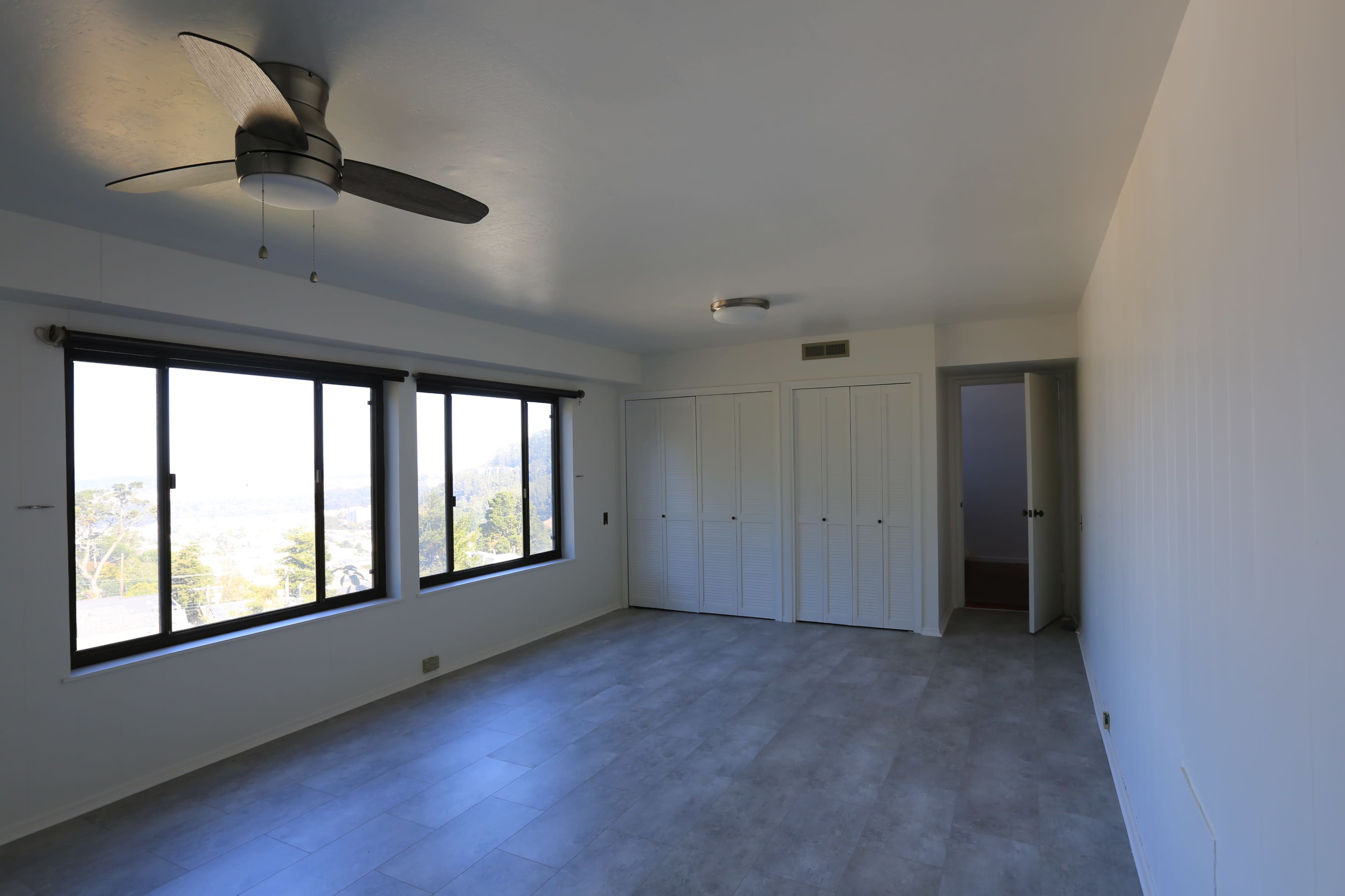 A spacious room features large windows, light-colored walls, and a ceiling fan, with two closets visible on one side.