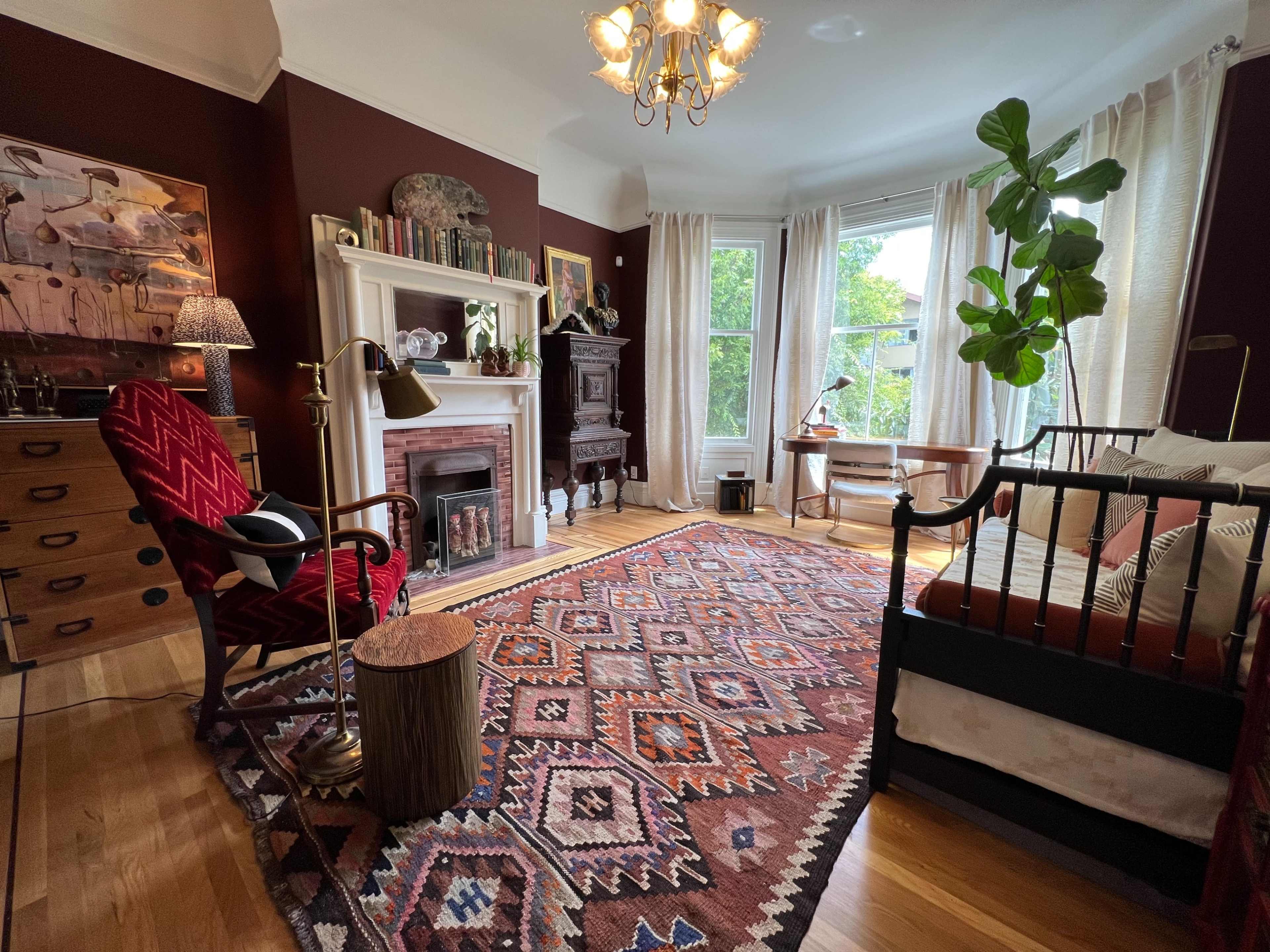 The room features a cozy living area with a patterned rug, a vintage armchair, a fireplace, and large windows providing natural light.