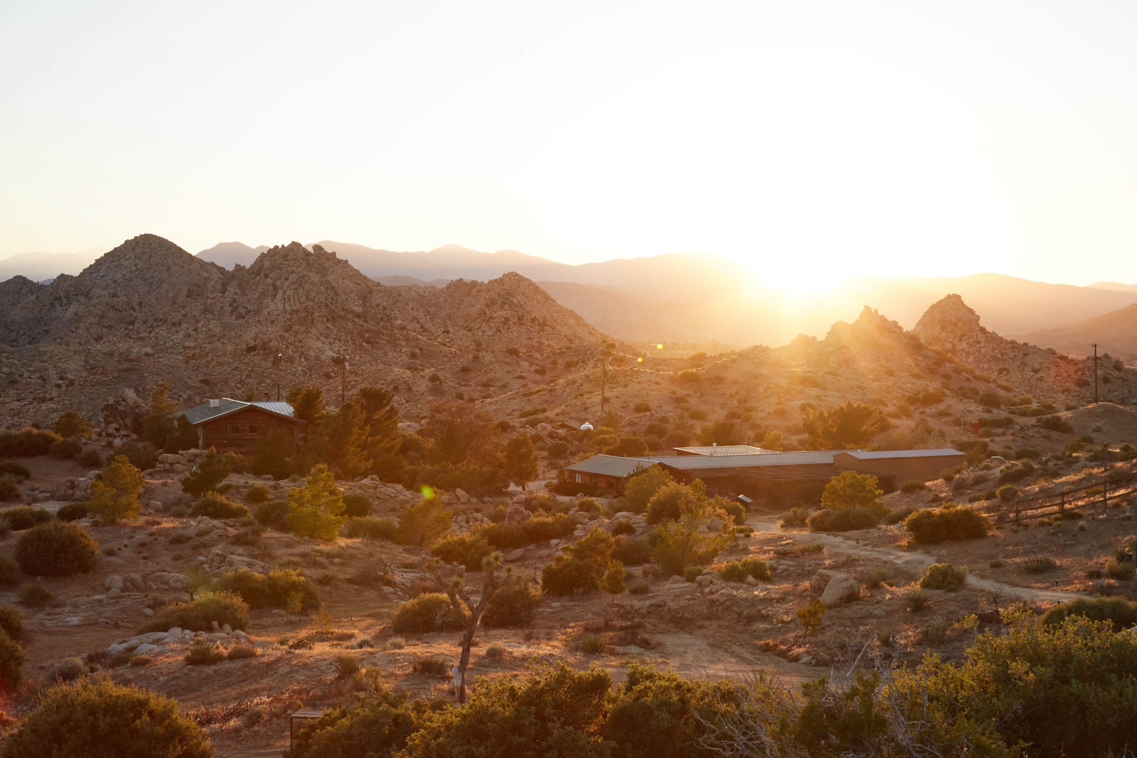 The sun rises over a rocky landscape dotted with sparse vegetation and a cluster of wooden buildings.