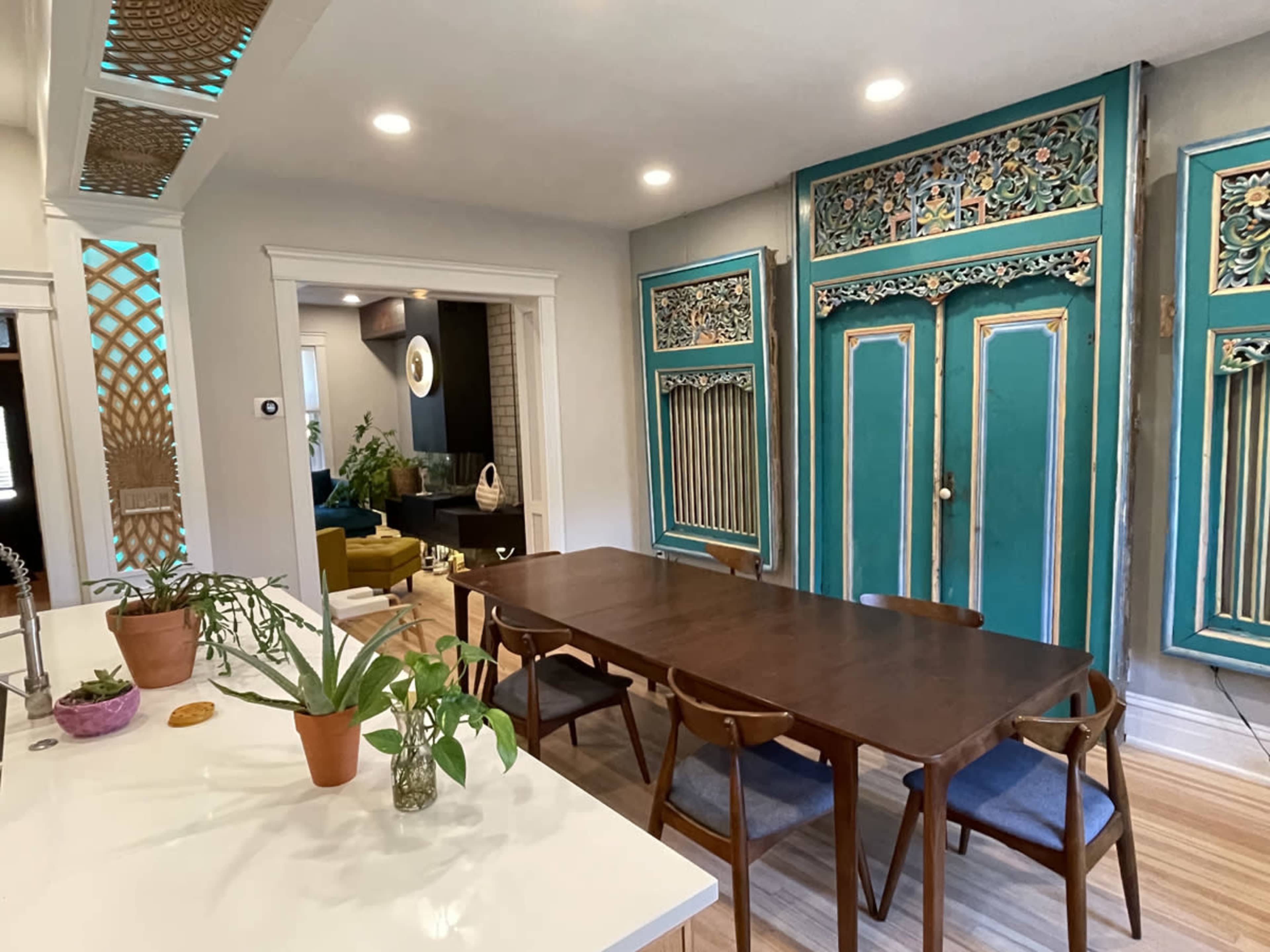 The image shows a dining area with a wooden table surrounded by chairs, set against a backdrop of ornate turquoise doors and plants on a countertop.