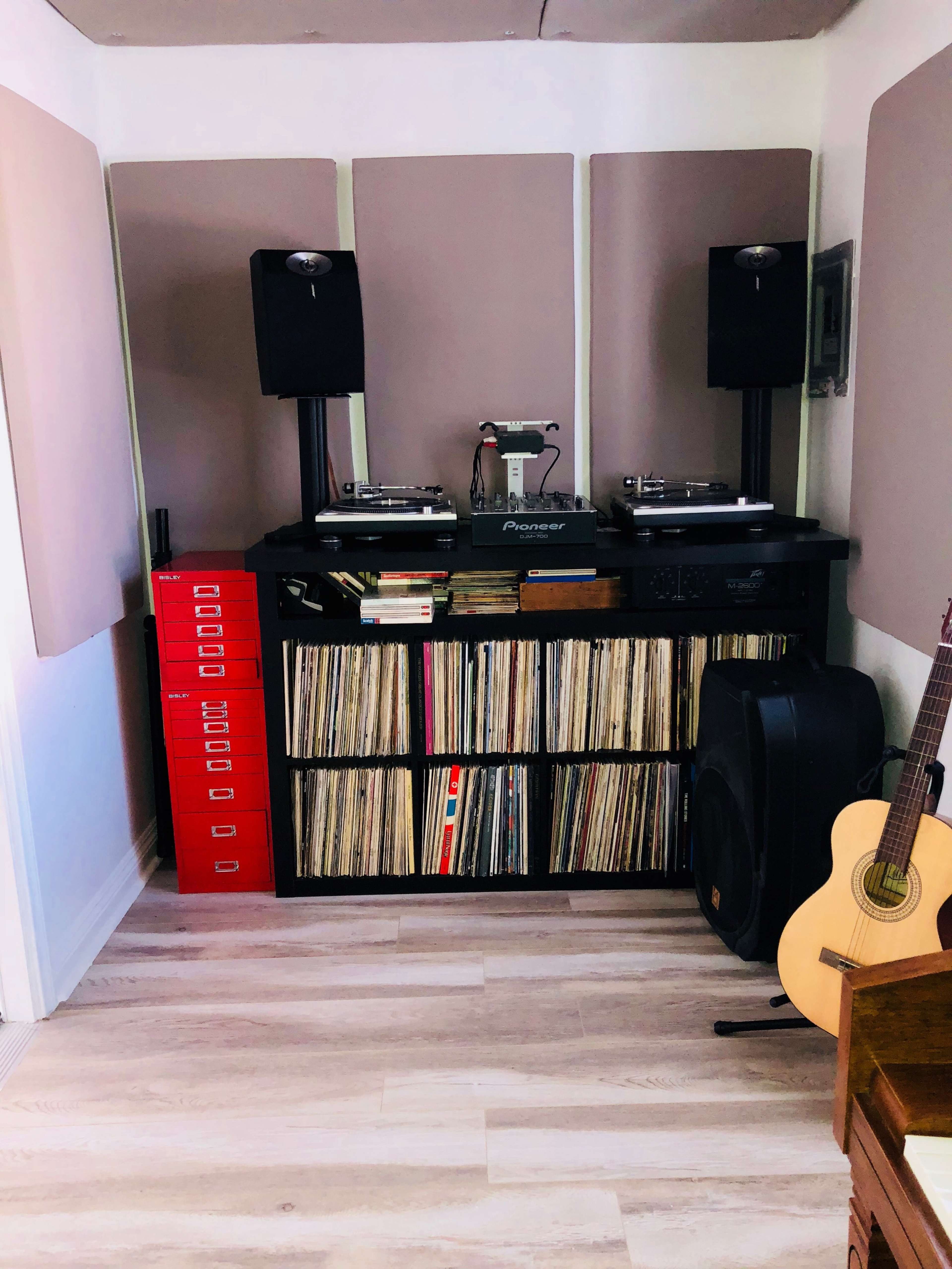 A music setup with two turntables on a black table, surrounded by speakers, a collection of vinyl records, and an acoustic guitar nearby.