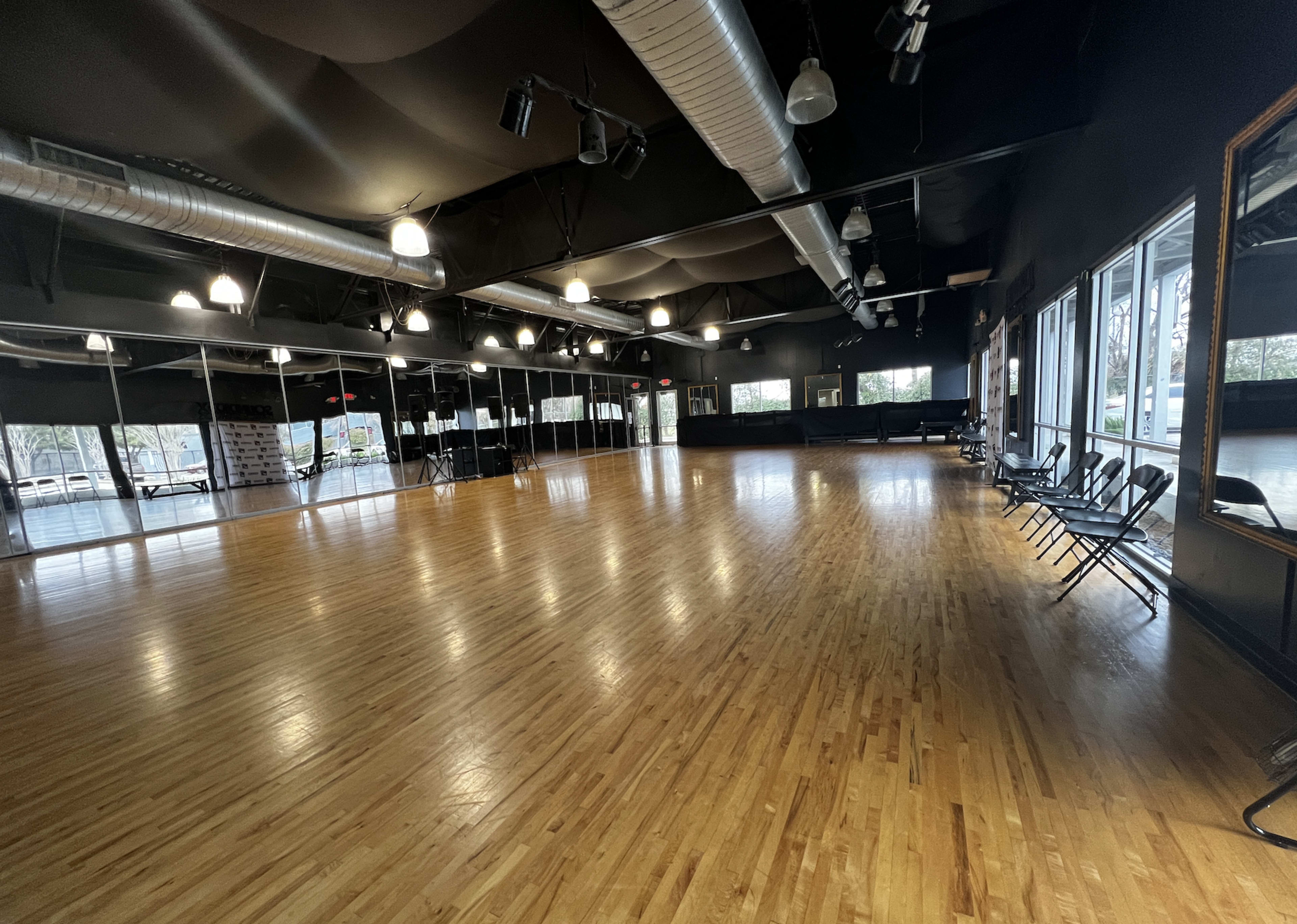 A spacious dance studio features polished wooden floors, black ceiling, and large windows allowing natural light.