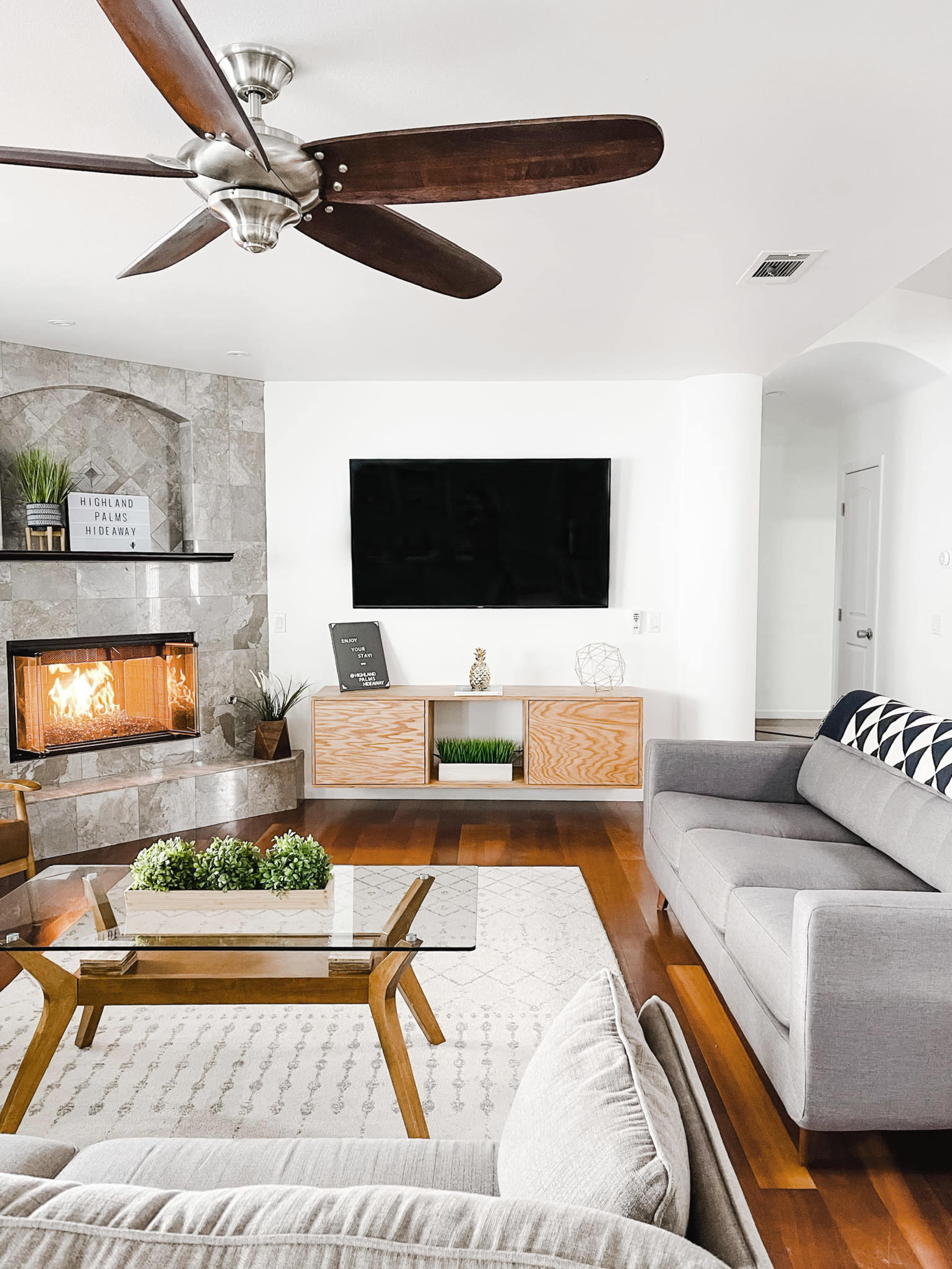 The living room features a modern design with a stone fireplace, a large television mounted on the wall, and various furniture pieces including a gray sofa and a wooden coffee table.